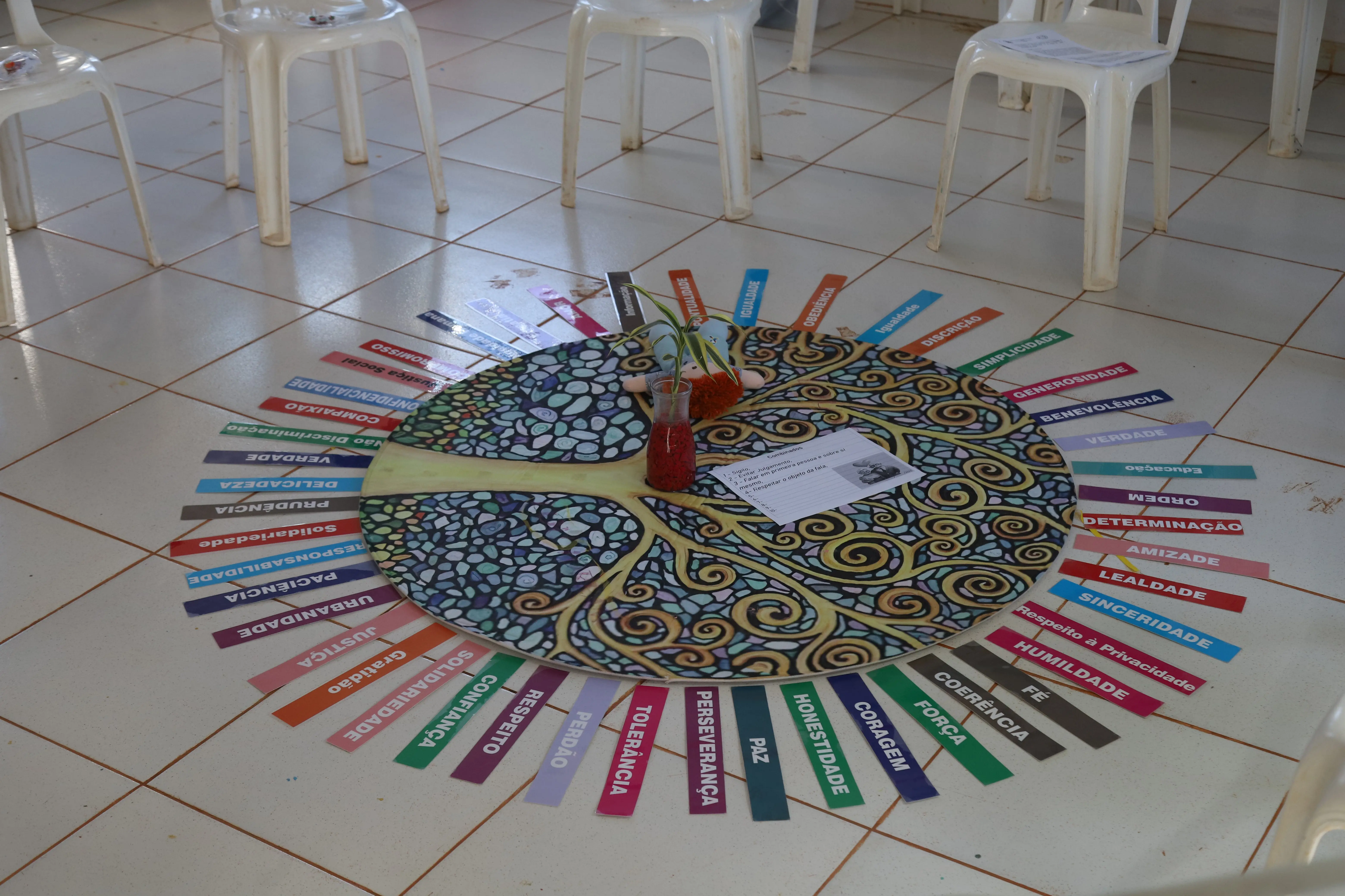 Mandala em forma de árvore ocupa o centro de uma roda de cadeiras vazias. Ao redor, palavras como “paz”, “coragem” e “confiança” representam valores humanos trabalhados na atividade do Círculo de Construção de Paz.