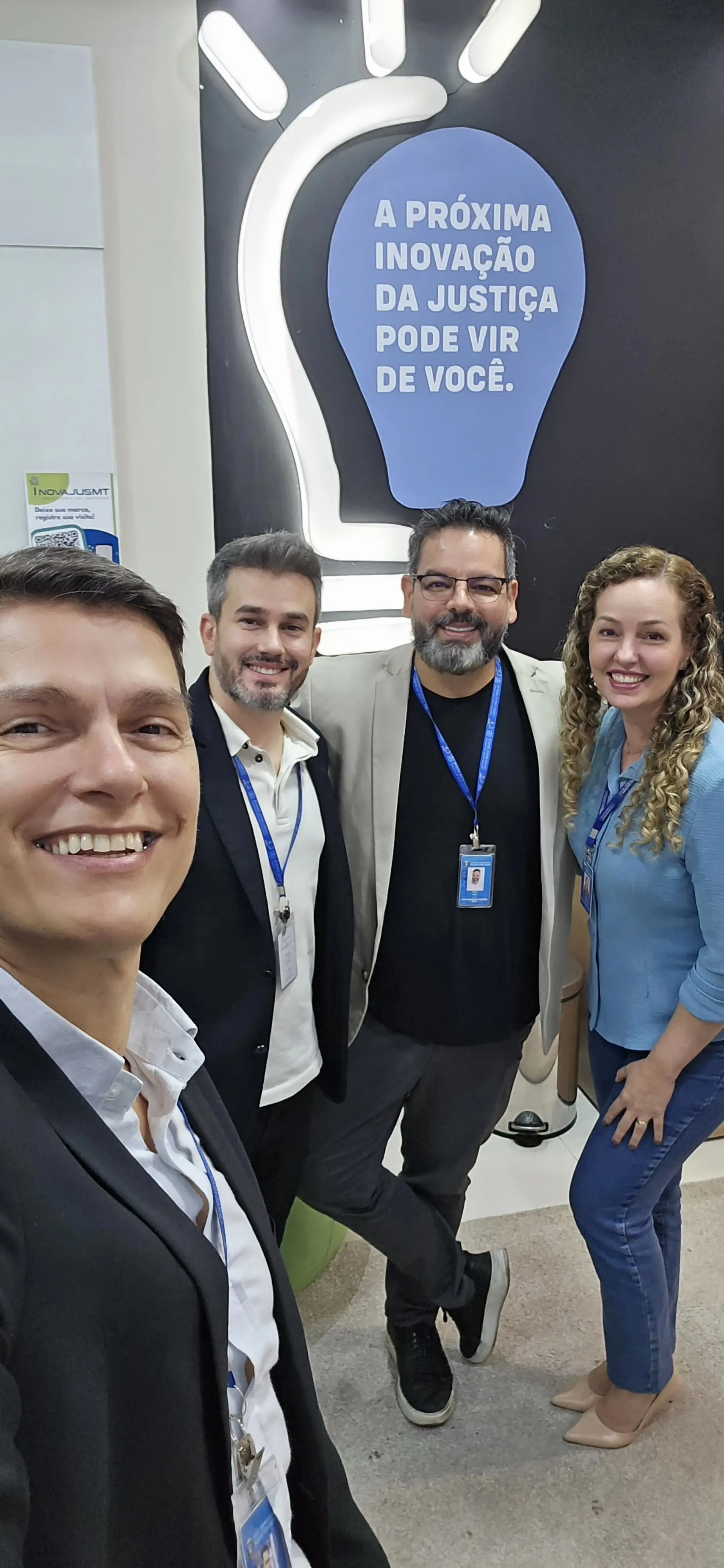 Quatro pessoas sorridentes posam para uma selfie. Três homens e uma mulher. Ao fundo, um painel com o desenho de uma lâmpada e a frase "A próxima inovação da Justiça pode vir de você."