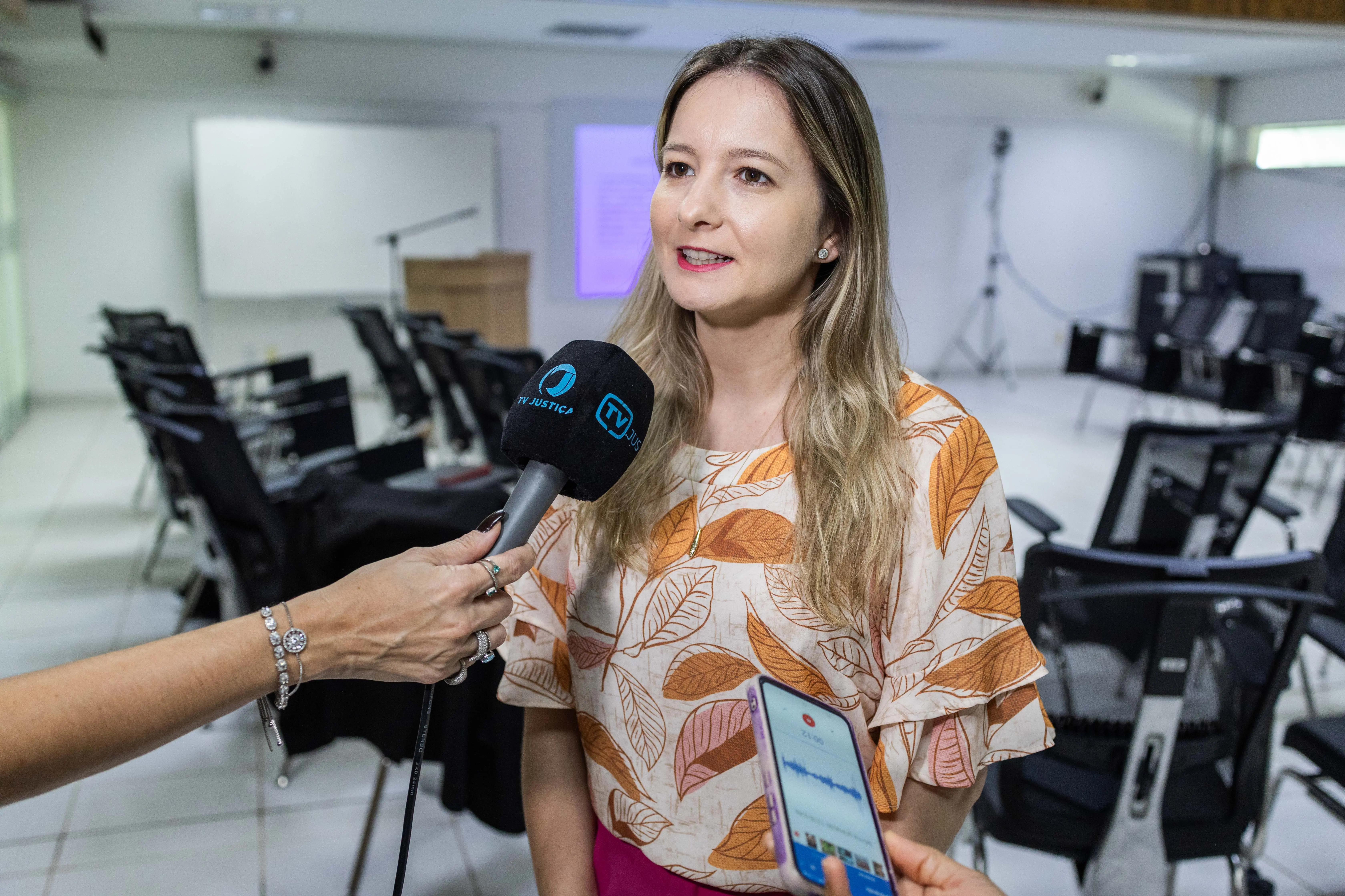 Juíza Djéssica Küntzer concede entrevista à TV.Jus. Ela é uma mulher branca, de cabelos longos e loiros, usando blusa com estampa de folhas em tons terrosos e batom vermelho. Ao fundo, é possível ver a sala de aula. 