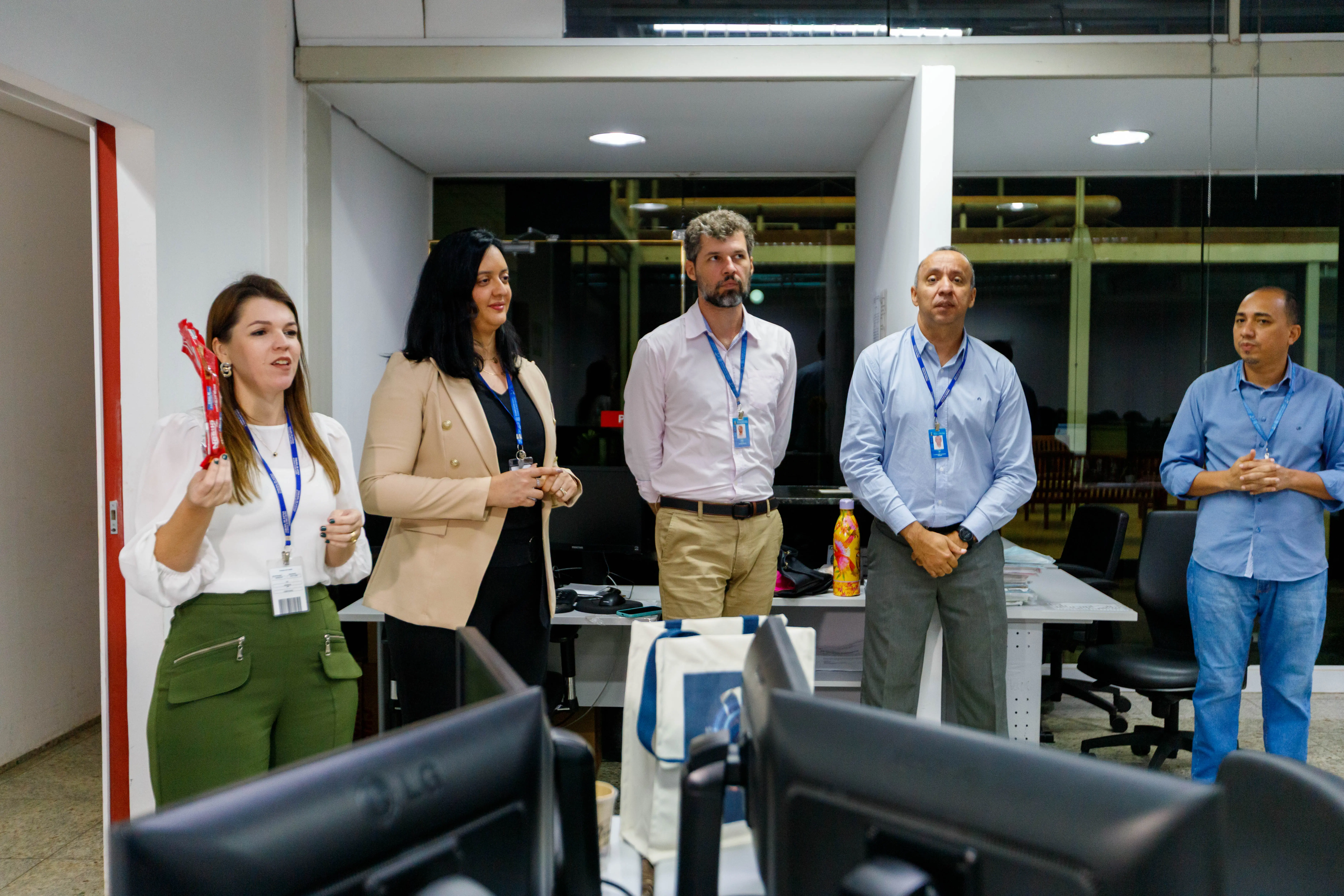 A imagem mostra um grupo de cinco pessoas em pé em um escritório. Na frente, à esquerda, uma mulher de blusa branca e calça verde segura uma embalagem plástica na mão direita enquanto fala.  Ao lado dela, outra mulher e três homens estão de pé, todos atentos. Eles vestem roupas sociais casuais. 