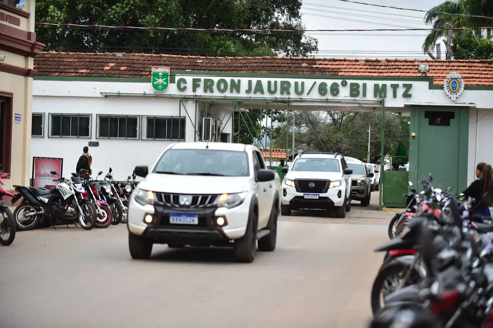 Imagem mostra veículos brancos saindo do quartel do Comando de Fronteira de Jauru (66º Batalhão), com placas visíveis e militares ao fundo. Motos estão estacionadas nas laterais e pessoas observam a movimentação.