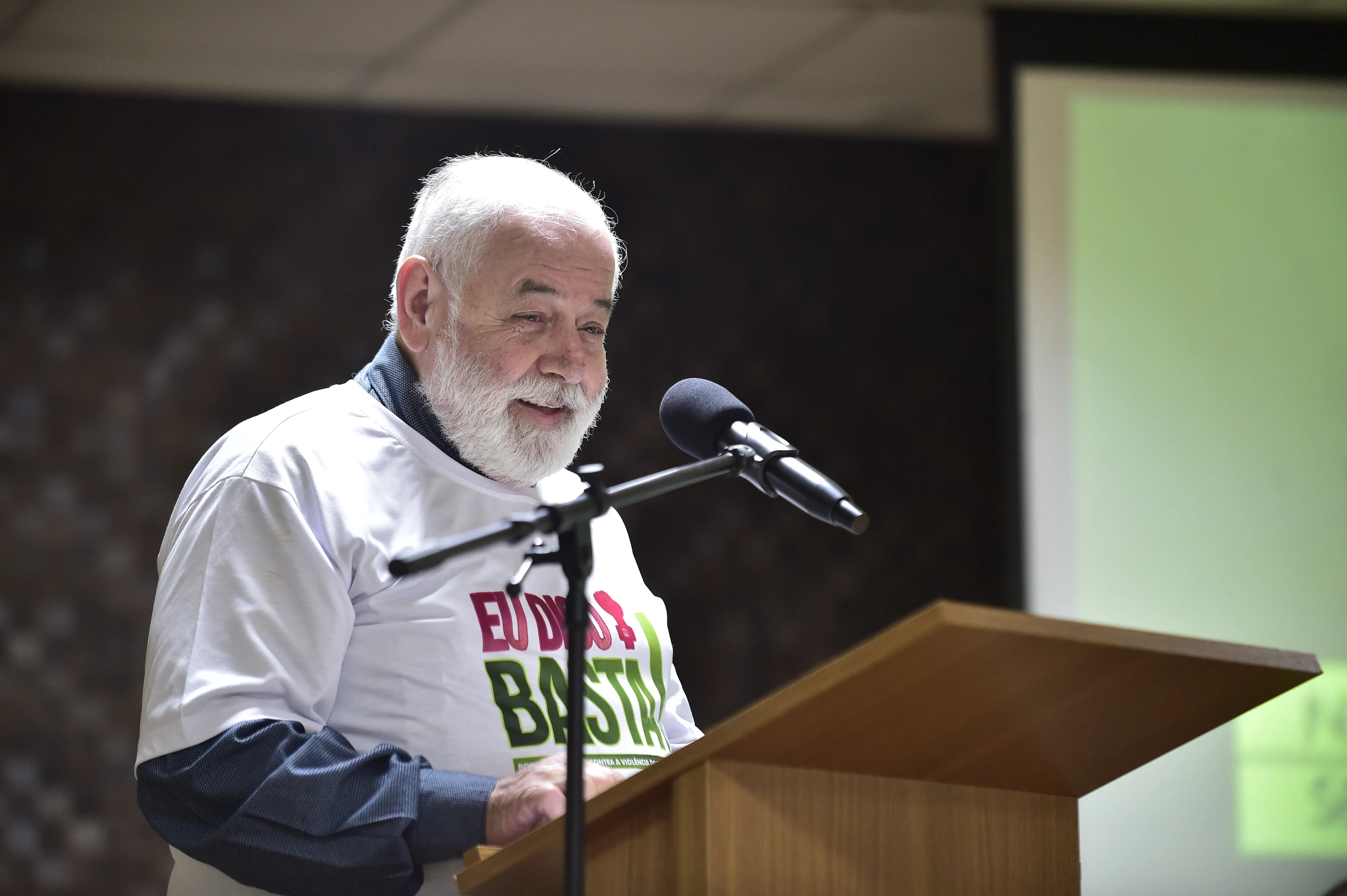 Foto do desembargador José Zuquim Nogueira falando no púlpito do auditório do TJMT. Ele é um senhor branco, de cabelos e barba brancos, usando a camiseta branca com a logo da campanha Eu Digo Basta. Ele está sorrindo.