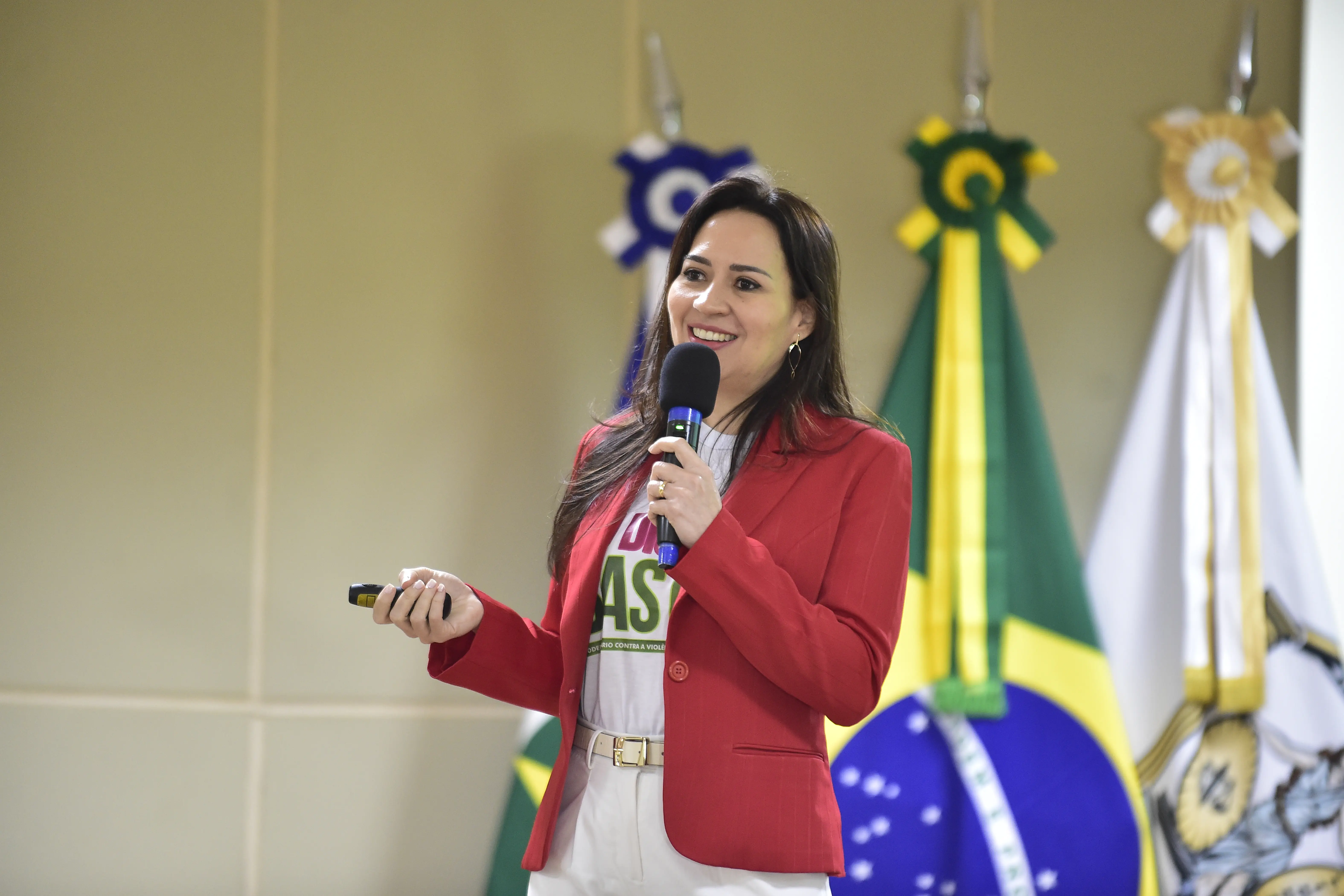 Juíza Tatyana Lopes proferindo palestra. Ela é uma mulher parda, de cabelos longos e castanhos, usando calça branca, camiseta da campanha Eu Digo Basta e terno vermelho.