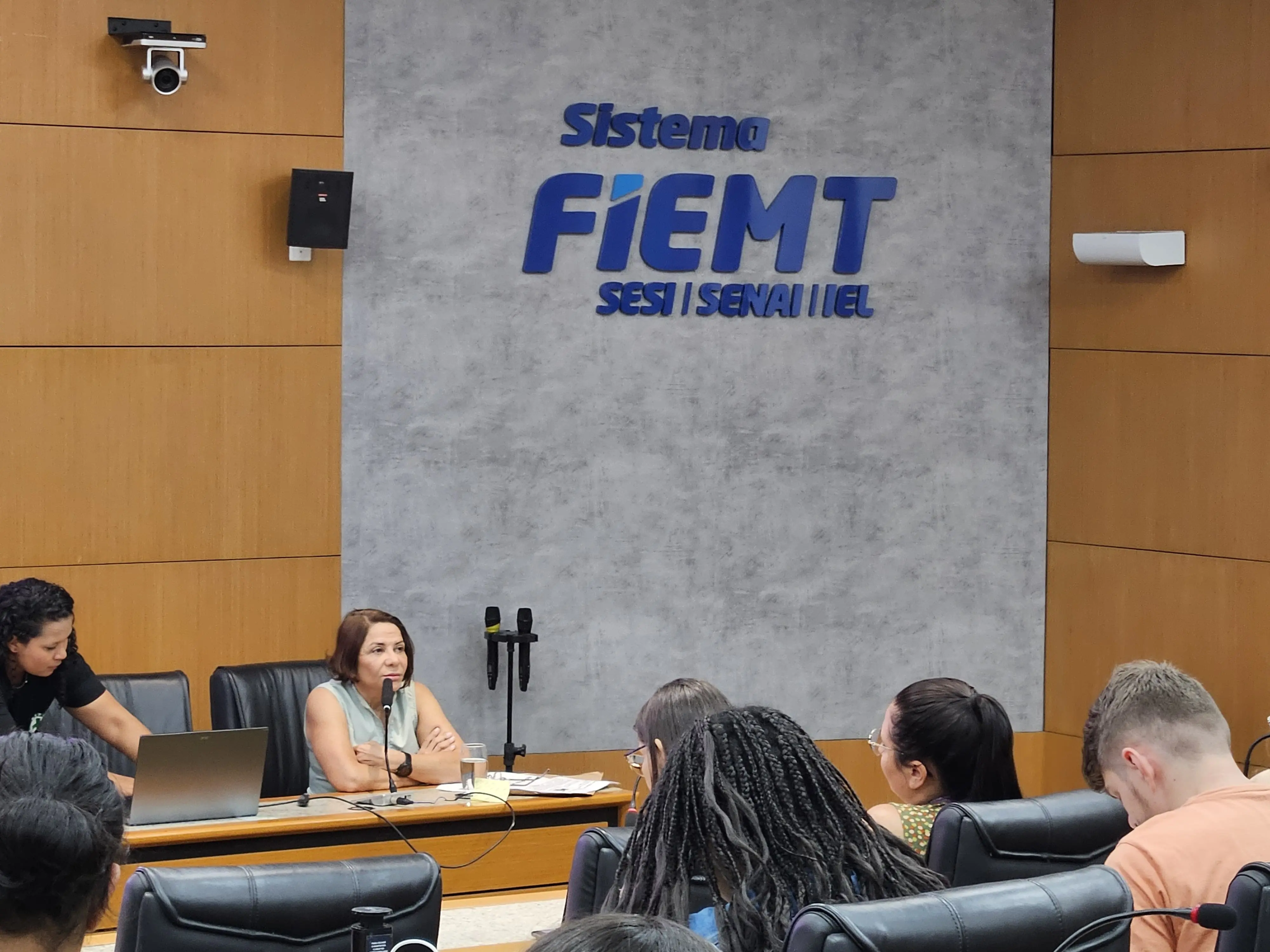 A imagem mostra um auditório ou sala de conferências com uma palestra em andamento. No centro, há uma mulher de cabelos castanhos, vestindo uma blusa branca, sentada em uma mesa de apresentação. Ela está olhando para a plateia, que ocupa as cadeiras em primeiro plano.