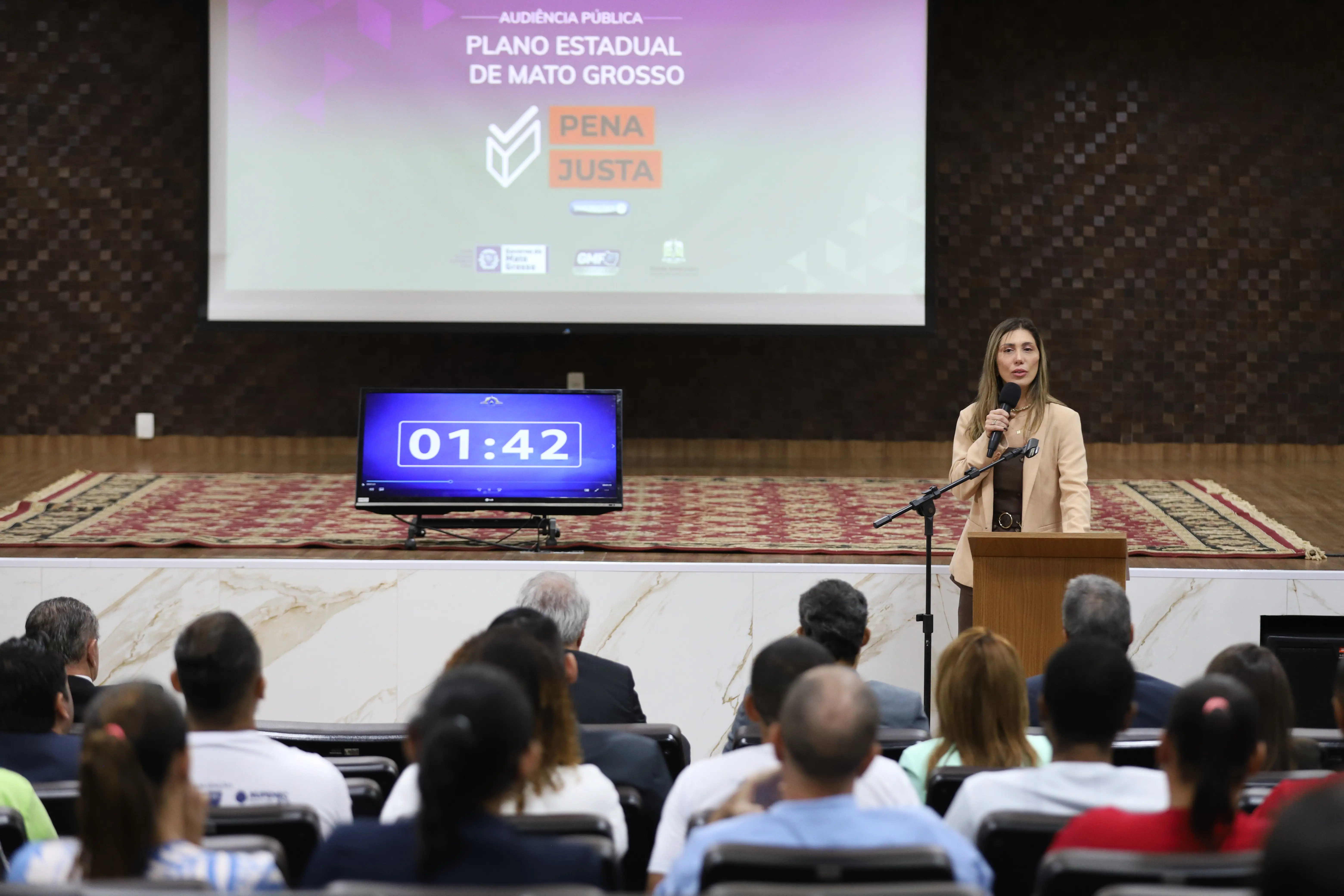 Uma mulher de cabelo liso e louro, usando um blazer bege, fala em um microfone sobre um púlpito de madeira. Atrás dela, um telão exibe "Audiência Pública Plano Estadual de Mato Grosso Pena Justa" e um relógio digital marca "01:42". Uma plateia assiste.