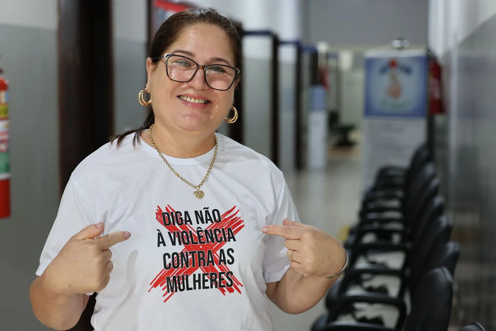 Mulher sorridente de óculos aponta para sua camiseta branca com os dizeres "DIGA NÃO À VIOLÊNCIA CONTRA AS MULHERES". Ela usa acessórios dourados e está em um corredor com cadeiras ao fundo.