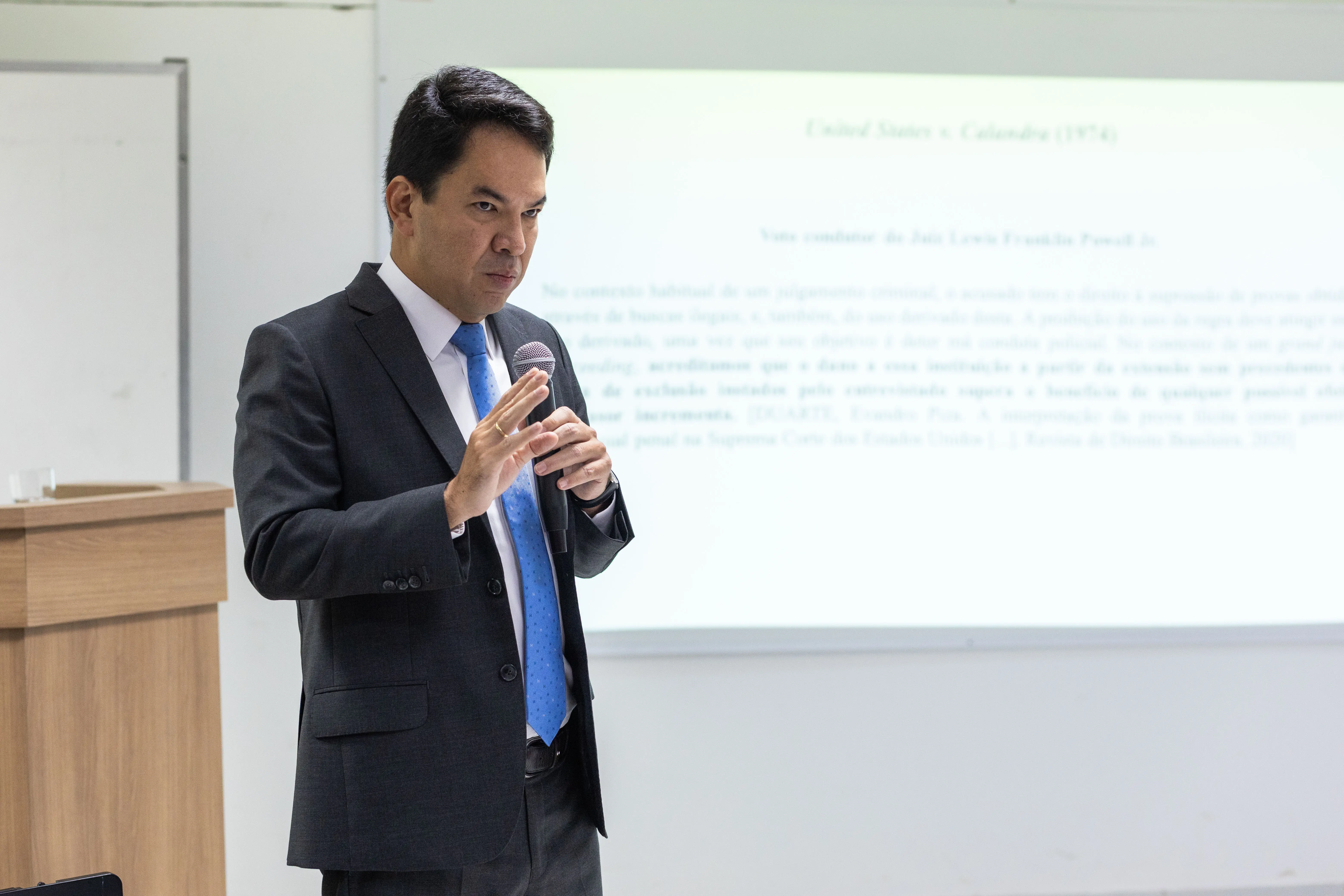  Hélio Nishiyama em pé, falando ao microfone e gesticulando. Ele é um homem pardo, de traços orientais, cabelos pretos e lisos, usando camisa branca, gravata azul clara e terno cinza escuro. Atrás dele, o conteúdo da aula está projetado na lousa branca. 