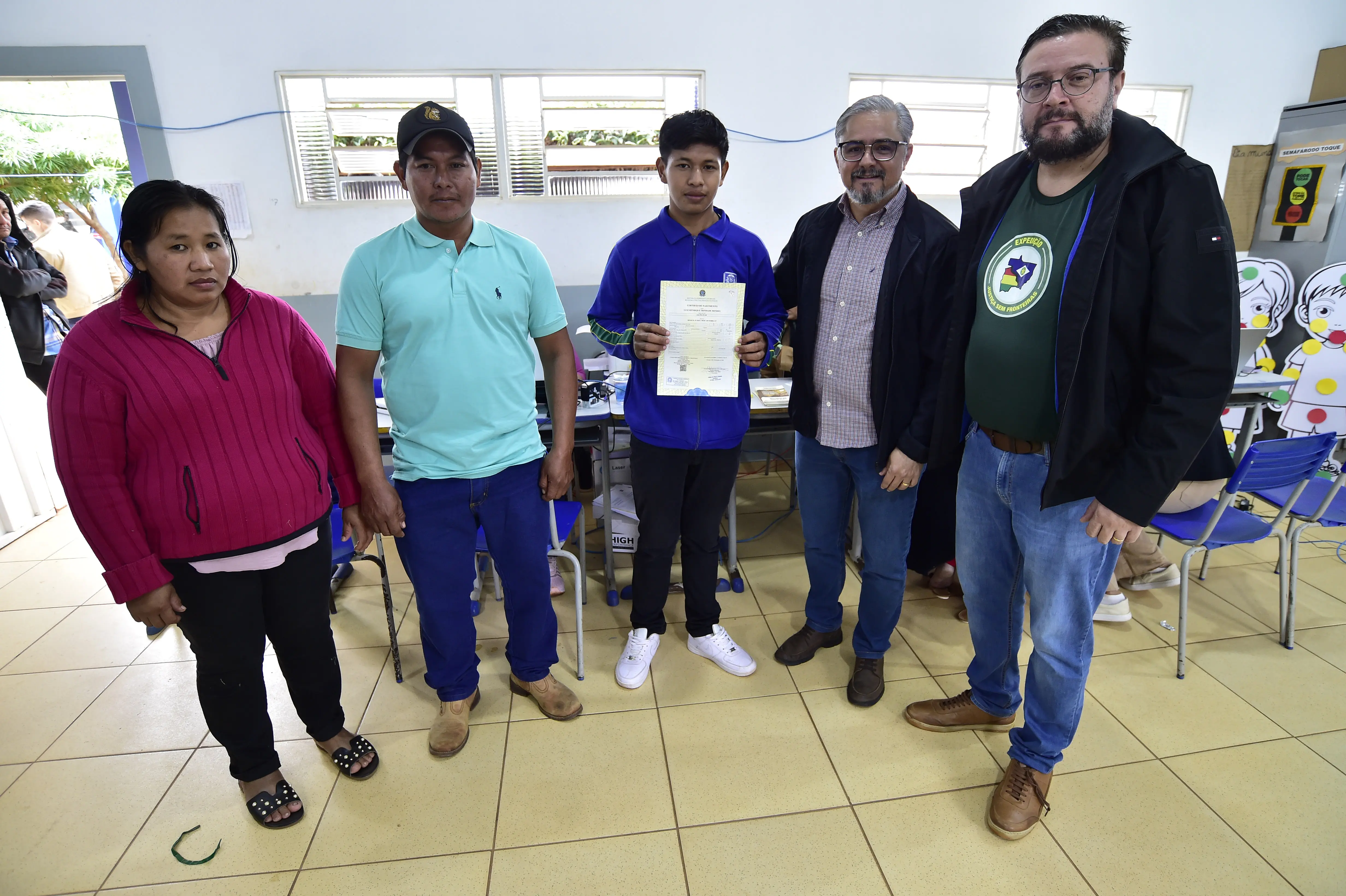 Cinco pessoas posam para foto em ambiente interno. Ao centro, um jovem segura uma certidão de nascimento. À esquerda, estão uma mulher de blusa rosa e um homem de boné e camisa verde. À direita, dois homens com barba e óculos, um com camisa xadrez e outro com camiseta verde do projeto.
