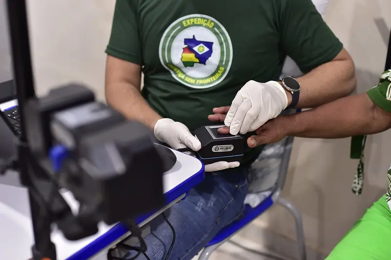 Foto em plano fechado que mostra servidor da Politec, usando camiseta verde da Justiça Sem Fronteiras, tirando biometria de um cidadão.