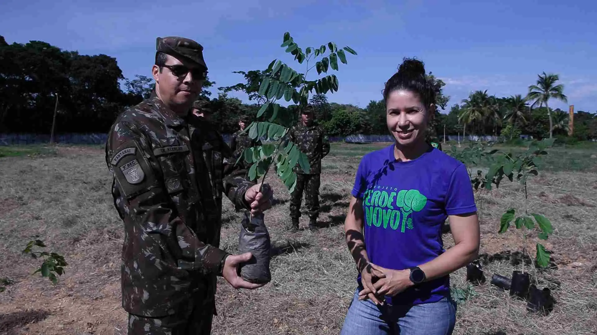 A foto mostra um homem em uniforme militar segurando uma muda de árvore em um saco plástico. Ao lado dele, uma mulher de camiseta azul com "PROJETO VERDE NOVO" e jeans sorri. Eles estão em um campo aberto, com outras pessoas e árvores plantadas no fundo