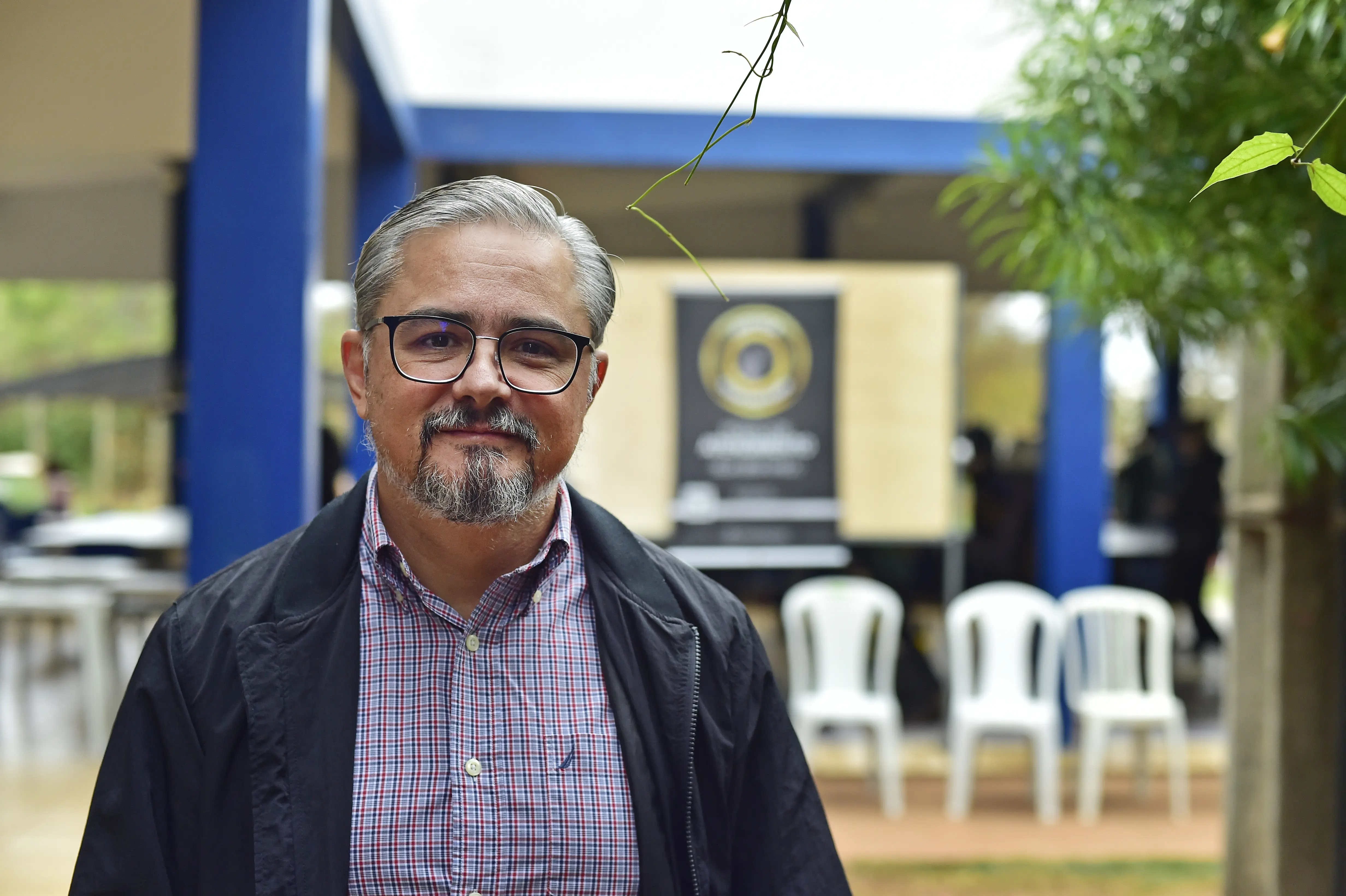 Homem de óculos, cabelo grisalho e barba aparece sorrindo levemente em primeiro plano. Ele veste camisa xadrez e jaqueta preta. Ao fundo, ambiente externo com cadeiras brancas e banner institucional desfocado. Há vegetação e colunas azuis no local.