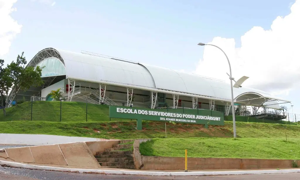 Fachada da Escola dos Servidores, localizada em um morro gramado. O prédio tem o teto semicircular e conta com um letreiro em concreto, fixo na grama.
