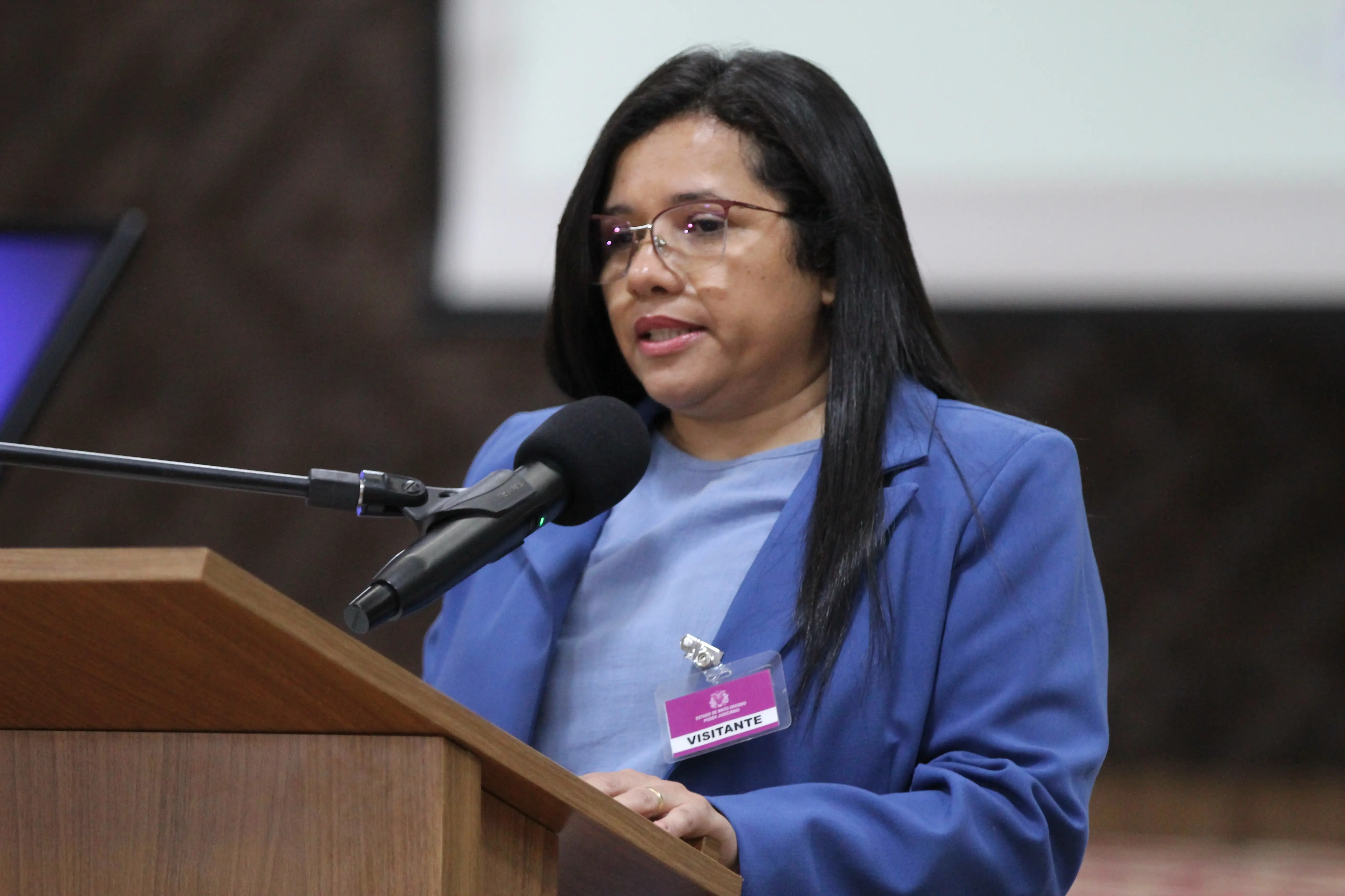 Eunice é mulher de cabelos pretos lisos e óculos, com um crachá de "VISITANTE", fala em um microfone sobre um púlpito de madeira. Ela usa um blazer azul e uma blusa azul clara.