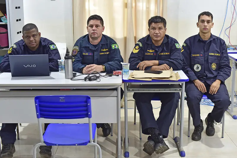 Quatro militares da Marinha posam para a foto. eles estão sentados, em uma sala de aula, uniformizados com fardas azul marinho.