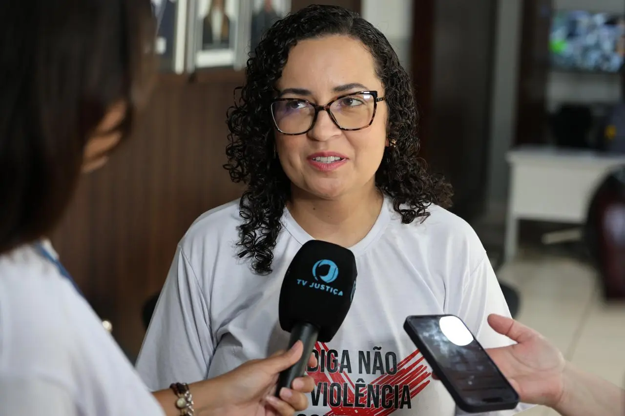 Uma mulher de cabelos cacheados escuros e óculos concede entrevista para microfones. Ela veste camiseta branca com a frase "DIGA NÃO À VIOLÊNCIA" em vermelho e preto, falando com expressão engajada diante das câmeras.