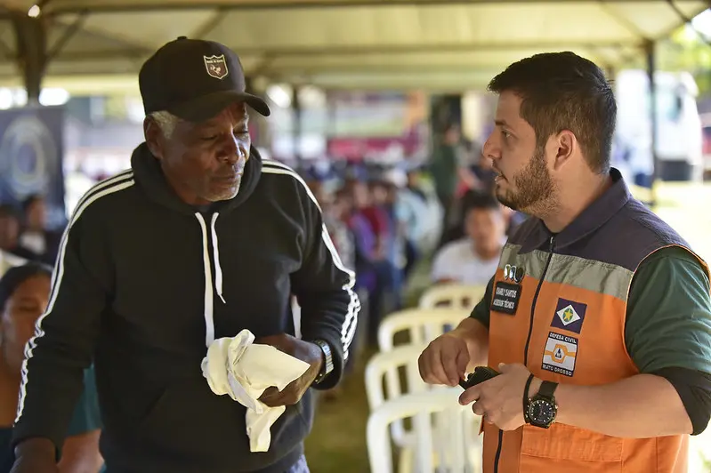 Servidor da Defesa Civil atende a um idoso, embaixo de uma tenda. Ele é um homem branco, usando colete azul e larajando.