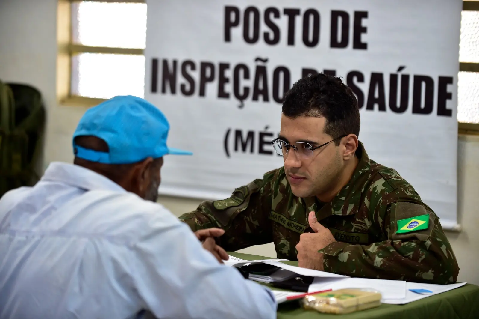 Militar do Exército atende homem idoso em posto de inspeção de saúde. Ambos estão sentados frente a frente em uma sala simples, com cartaz informativo ao fundo indicando o serviço médico.