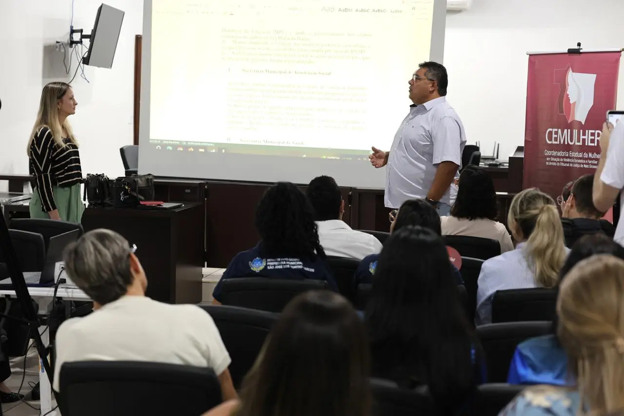 Um homem de camisa branca apresenta para uma plateia sentada, gesticulando enquanto fala. Ao seu lado, uma mulher loira de blusa preta listrada observa. Ao fundo, há um banner da CEMULHER e uma tela de projeção com documento.