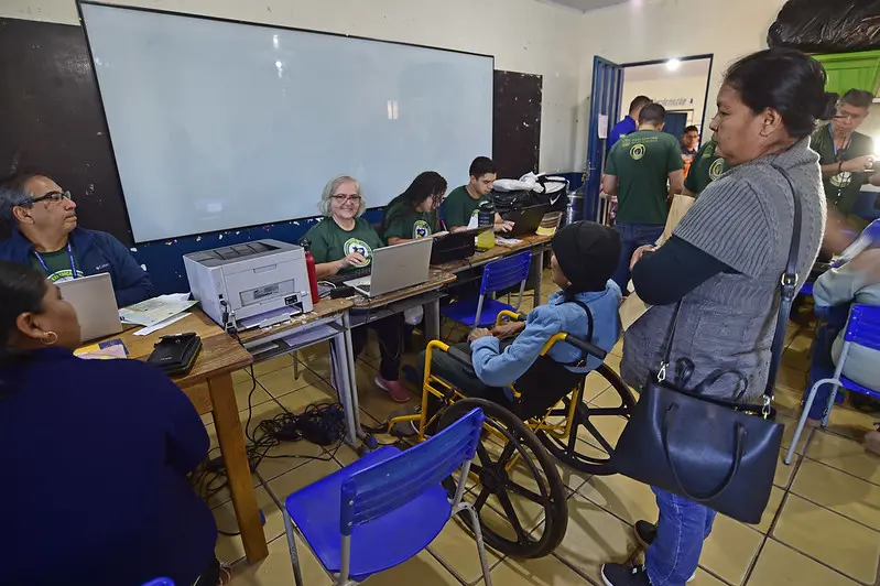 Servidores da Receita Federal e da Polícia Federal realizam atendimentos aos cidadãos, em uma sala de aula. Eles usam a camiseta verde musgo da Justiça Sem Fronteiras.