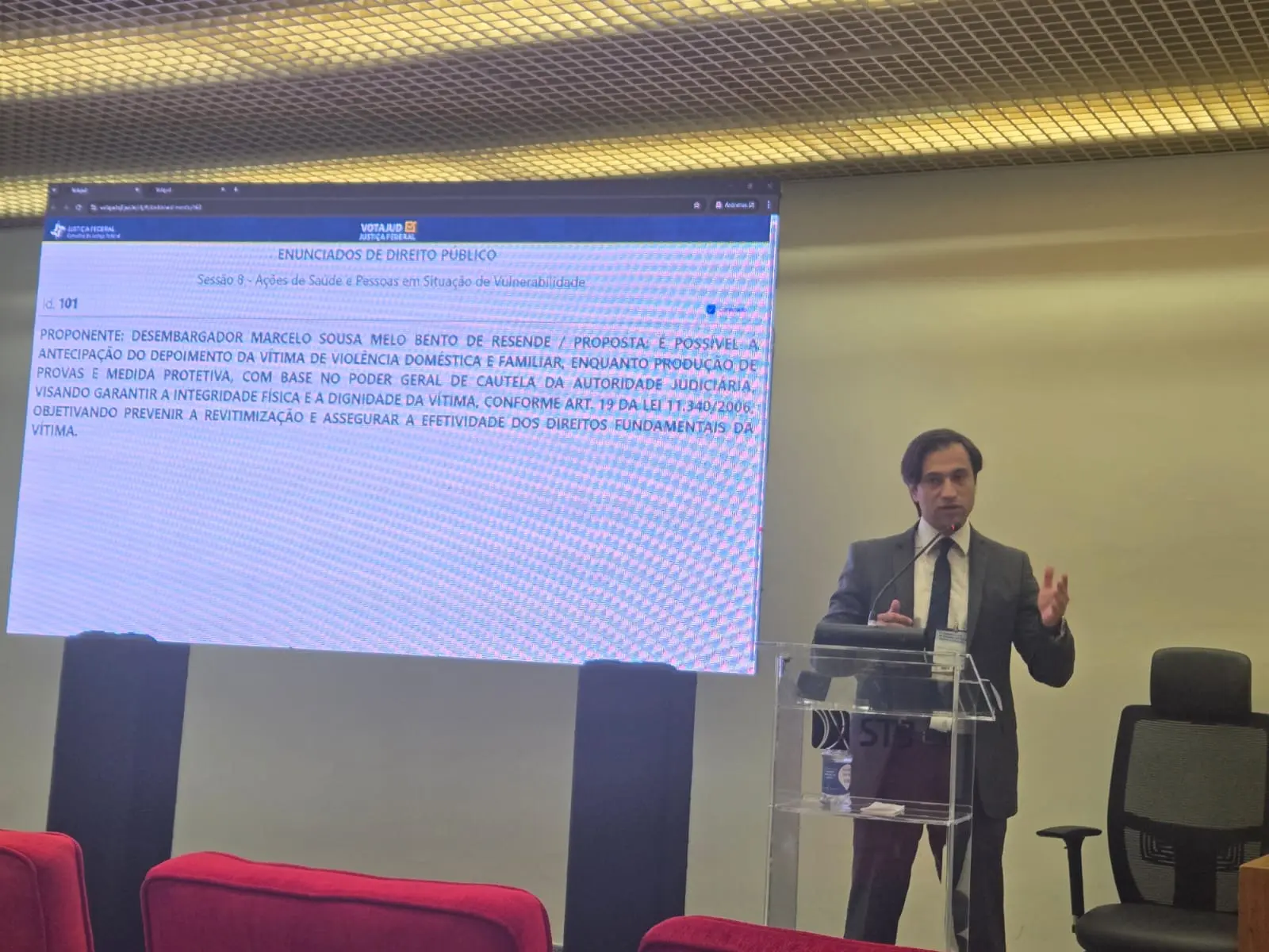 Juiz Marcelo Resende fala em púlpito do STJ, diante de telão que exibe enunciado jurídico sobre violência doméstica e medidas protetivas. Ele propõe antecipação de depoimento para garantir segurança da vítima.