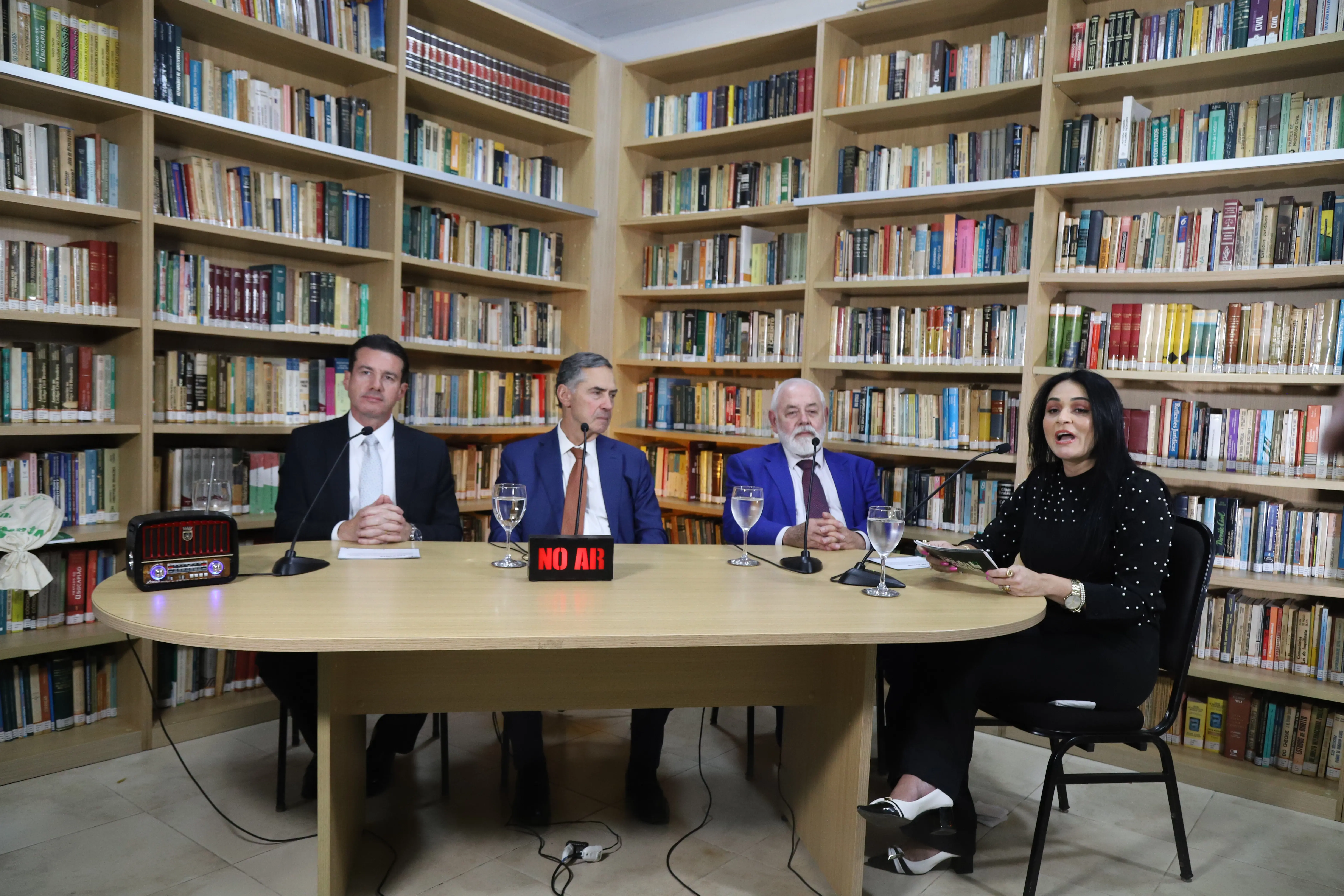 Presidente do TJMT, desembargador José Zuqui, desembargador Rodrigo Curvo estão ao lado do Ministro Barroso e a apresentadora estão atentos durante a gravação. Livros preenchem as estantes ao fundo, e a mesa central destaca a luz vermelha da placa “NO AR”, reforçando o ambiente formal.