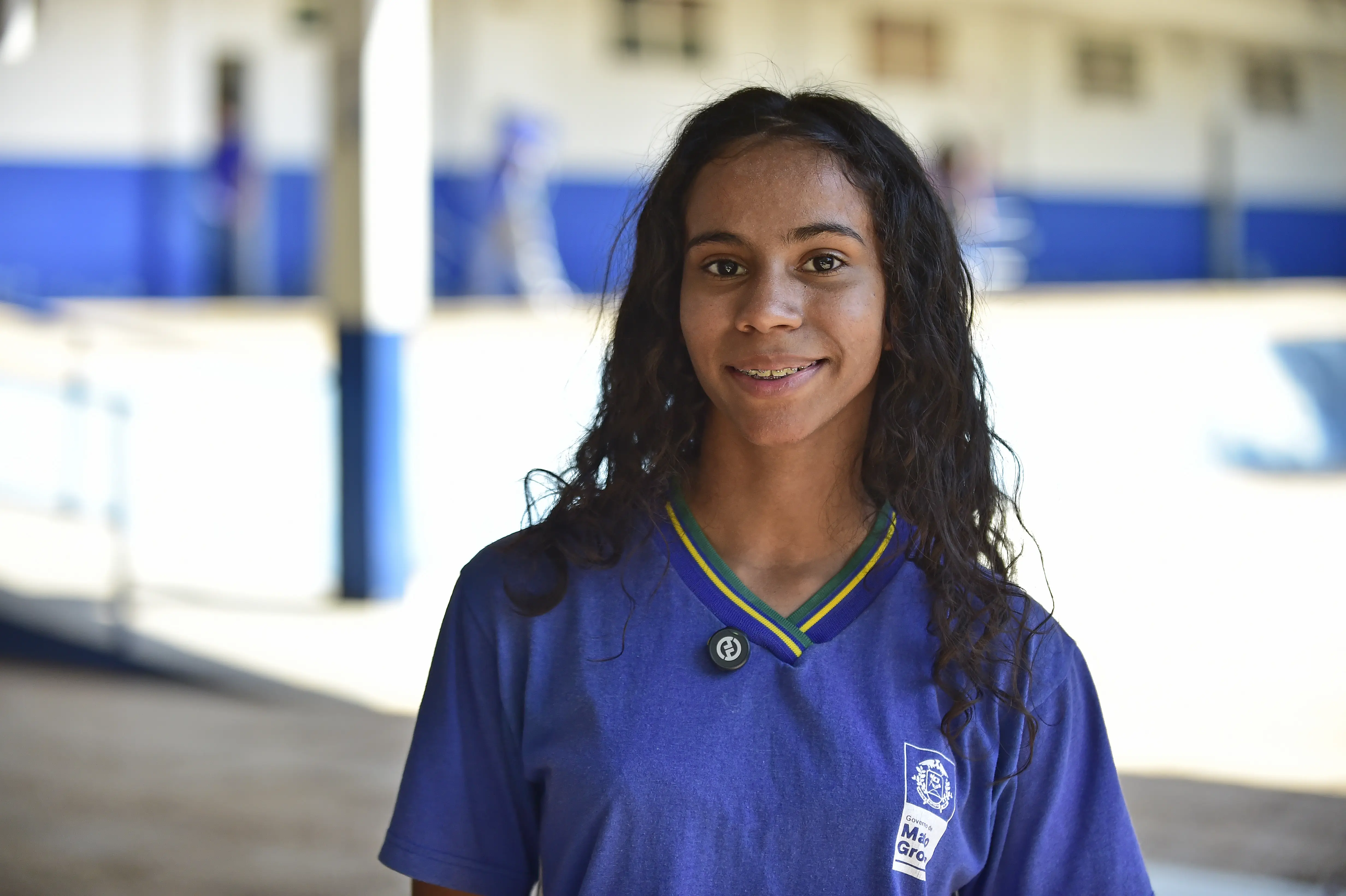 Caroline Souza Santos, 17 anos, está sorrindo, tem cabelo cacheado escuro e veste uma camiseta azul com detalhes em verde e amarelo. Ao fundo, um pátio escolar com colunas e paredes azuis e brancas.