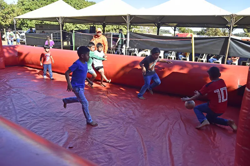 Crianças brincam de futebol em um campo inflável, no pátio de uma escola. Um bombeiro militar atua como árbitro do jogo.