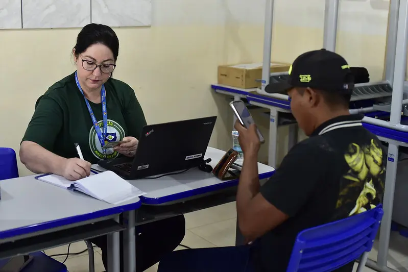 Funcionária da Caixa Econômica Federal atende a um homem, numa sala de aula. ela é uma mulher branca, de cabelos pretos, usando óculos de grau, crachá e camiseta verde da Justiça Sem Fronteiras.