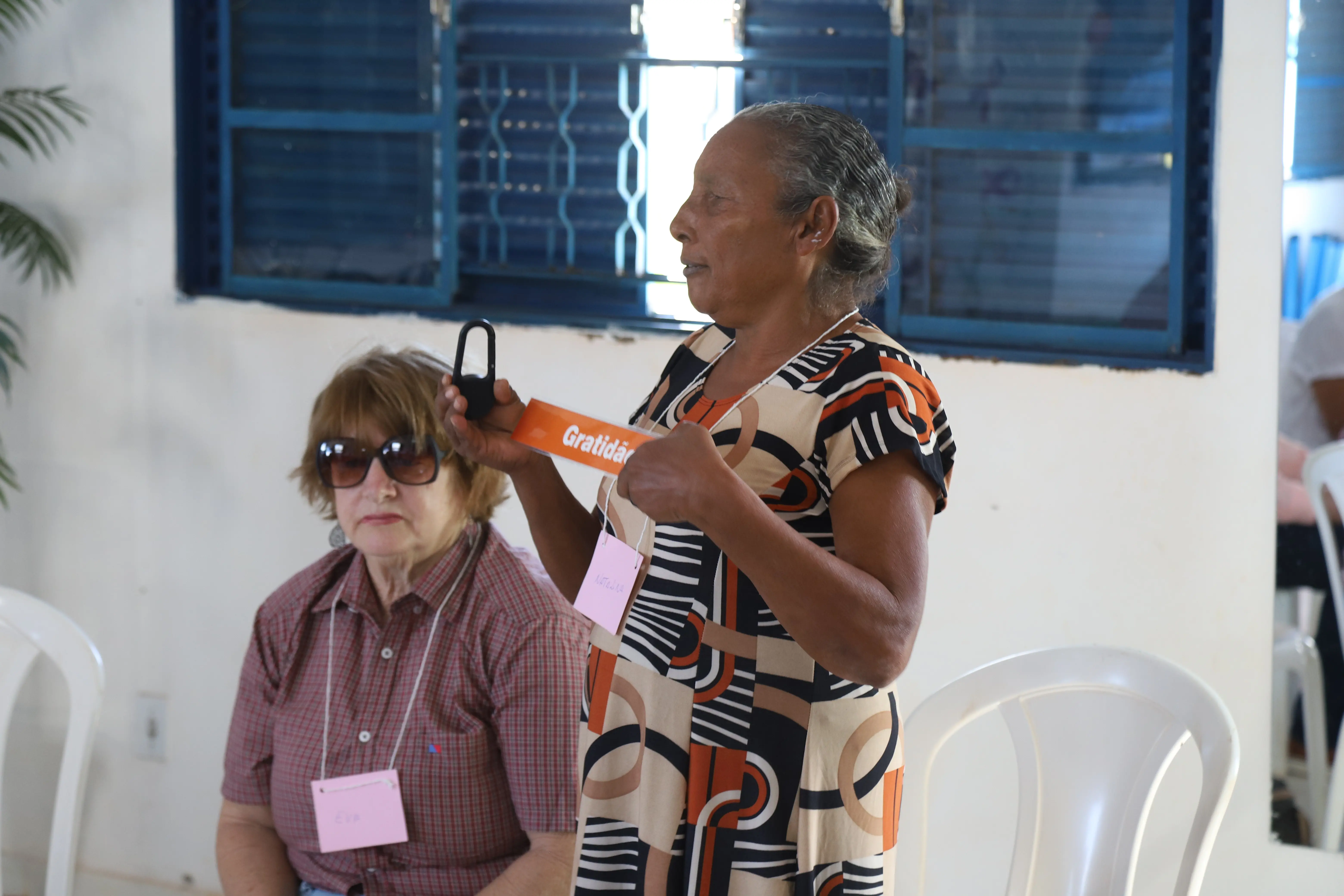 Mulher em pé compartilha a palavra “Gratidão” segurando também uma bússola. Com crachá “NATÁLIA”, ela participa ativamente da roda, enquanto demais integrantes acompanham sentadas e atentas.