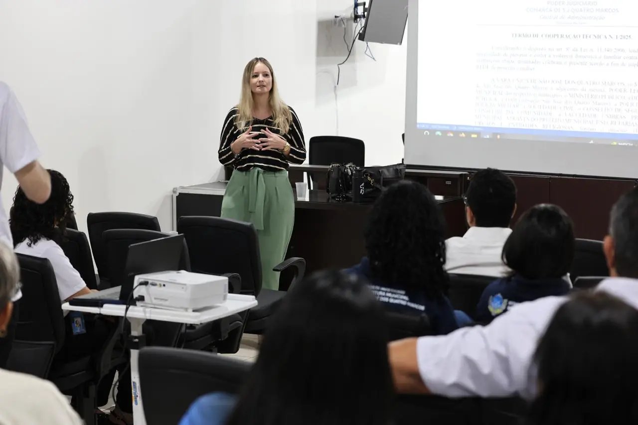 Uma mulher de cabelos loiros apresenta para uma audiência em uma sala de reuniões. Ela veste blusa preta listrada e calça verde, segurando um celular. Atrás dela, uma tela de projeção exibe um documento sobre cooperação técnica.