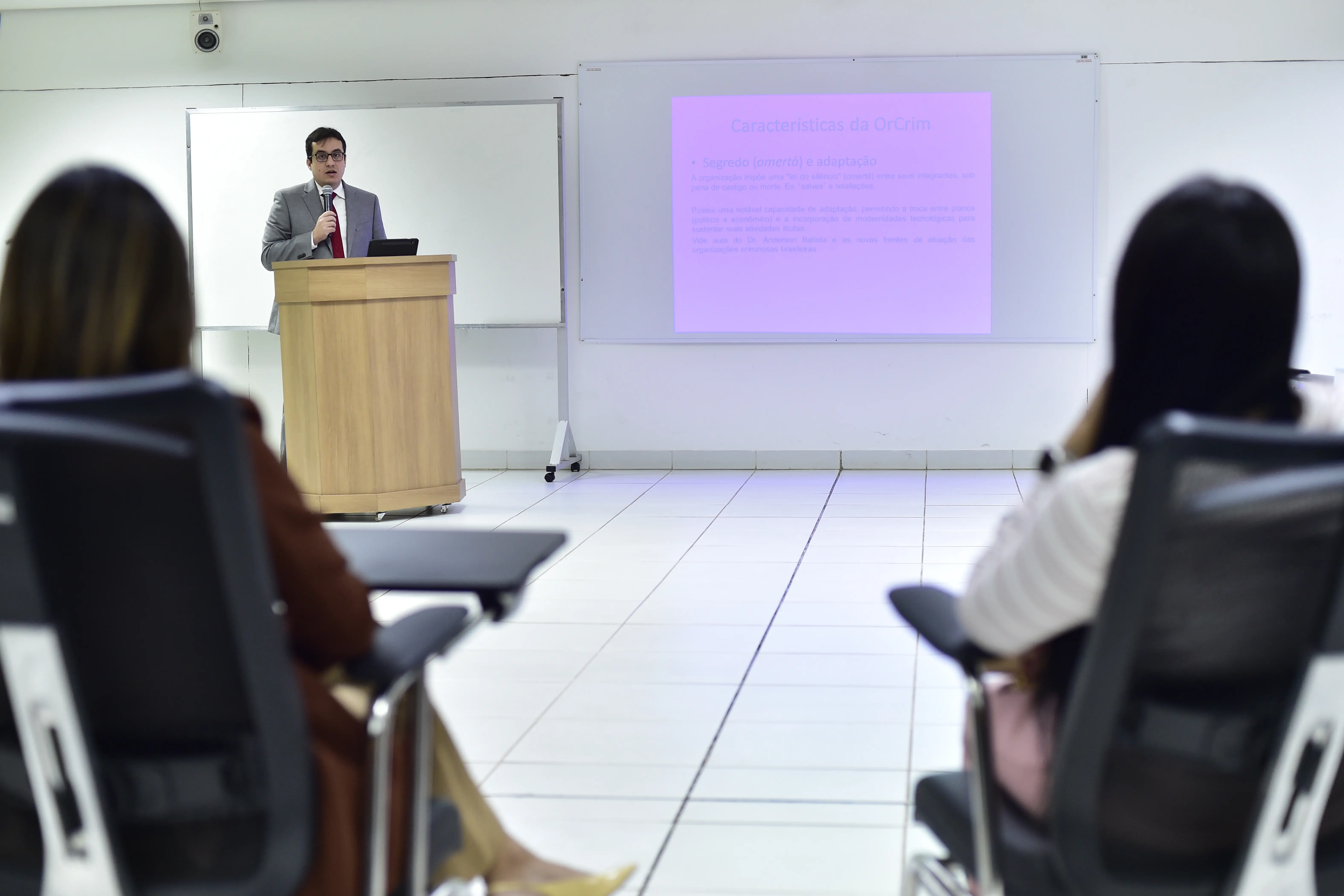 Homem de terno fala ao microfone atrás de um púlpito, em sala com cadeiras e projeção sobre organizações criminosas. À frente, duas mulheres sentadas assistem à apresentação com atenção.