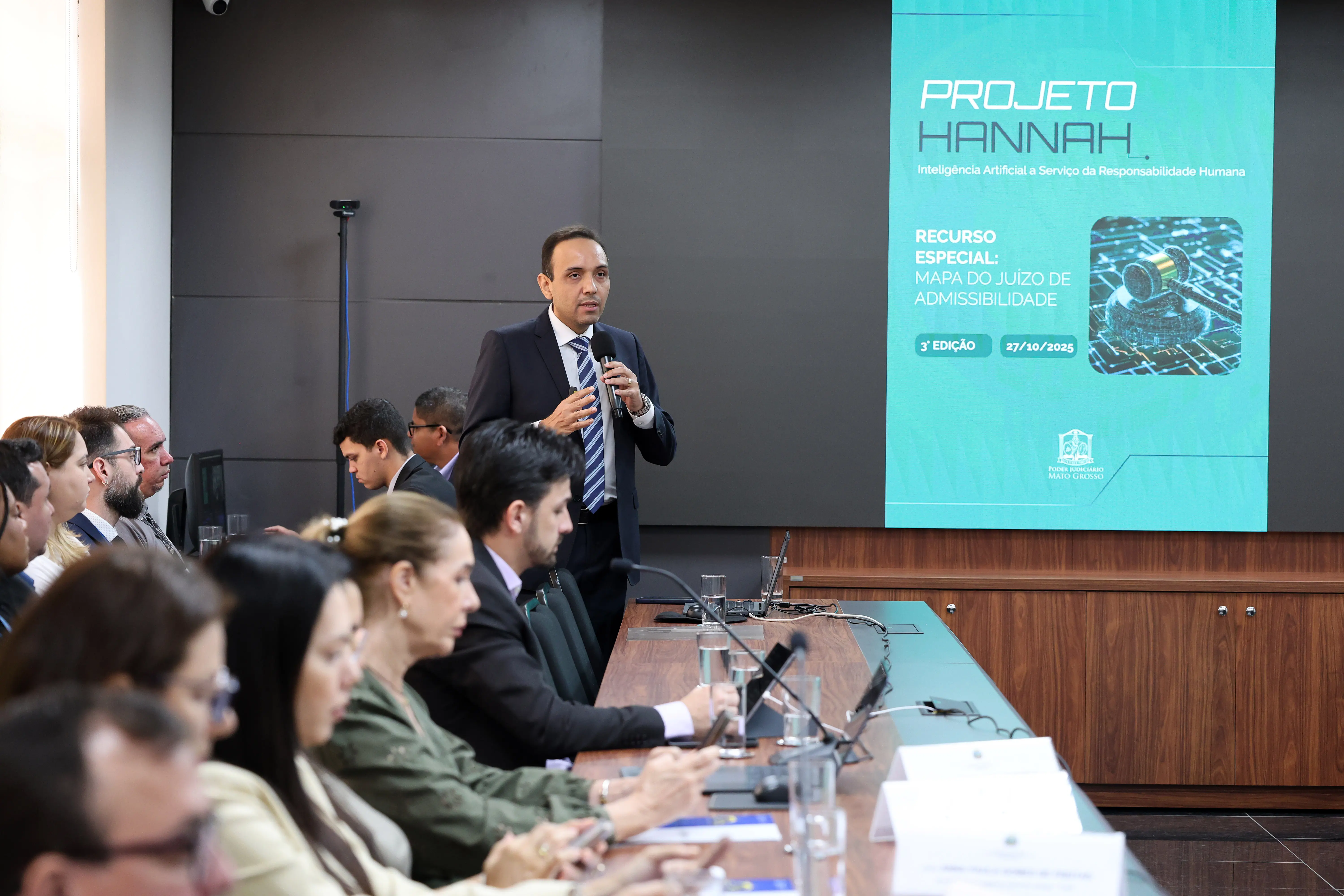 Plano aberto de sala de reunião em formato de "U". Um servidor apresenta slides sobre "Ferramentas / Stack Tecnológico" em uma tela ao fundo para diversos participantes atentos