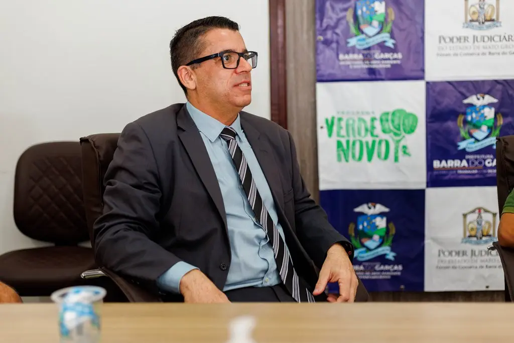 Juiz de Barra do Garças, Michell Lotfi, sentado, falando. Ele é um homem de pele clara, usando óculos de grau, camisa azul, terno cinza escuro e gravata listrada preta e cinza. Atrás dele há um painel com as logomarcas do programa Verde Novo, do Poder Judiciário e brasão de Barra do Garças.