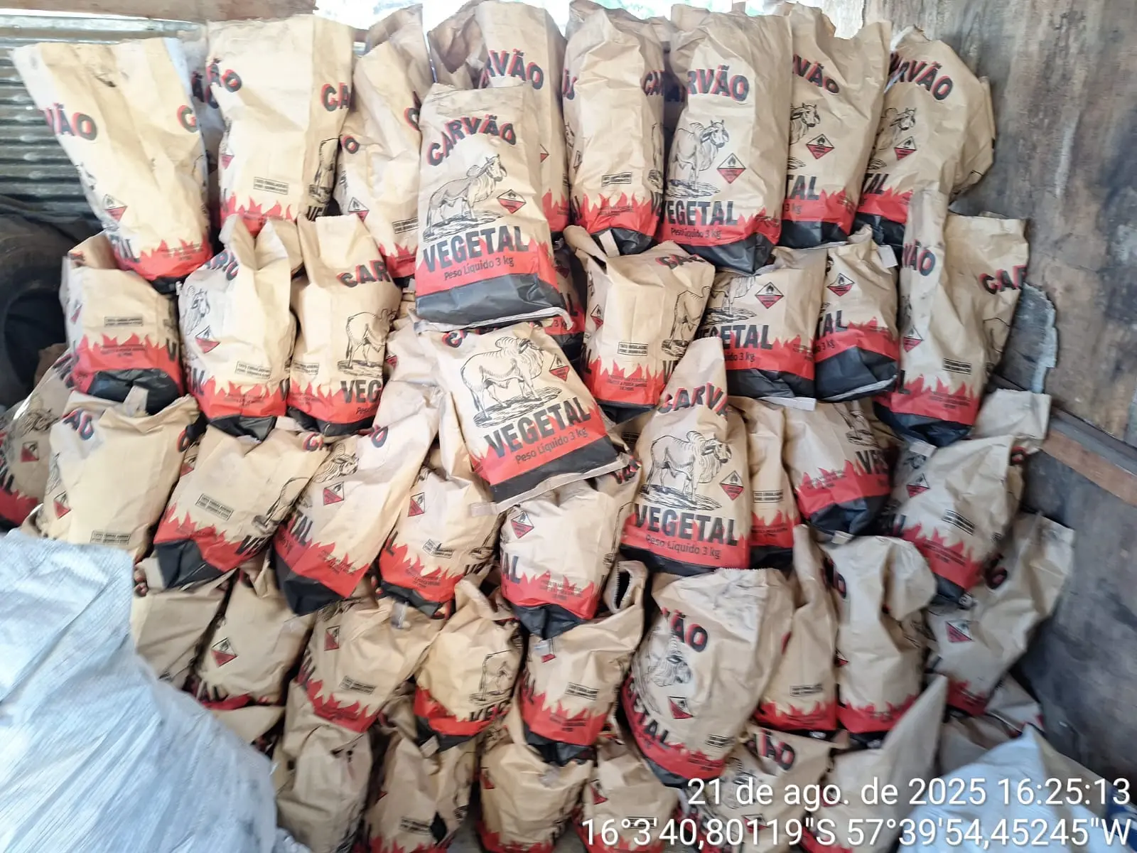 Esta foto é um close da pilha de sacos de carvão. Os sacos são de papel pardo e têm a inscrição "CARVÃO VEGETAL Peso Líquido 3 kg" e o desenho de um boi. 