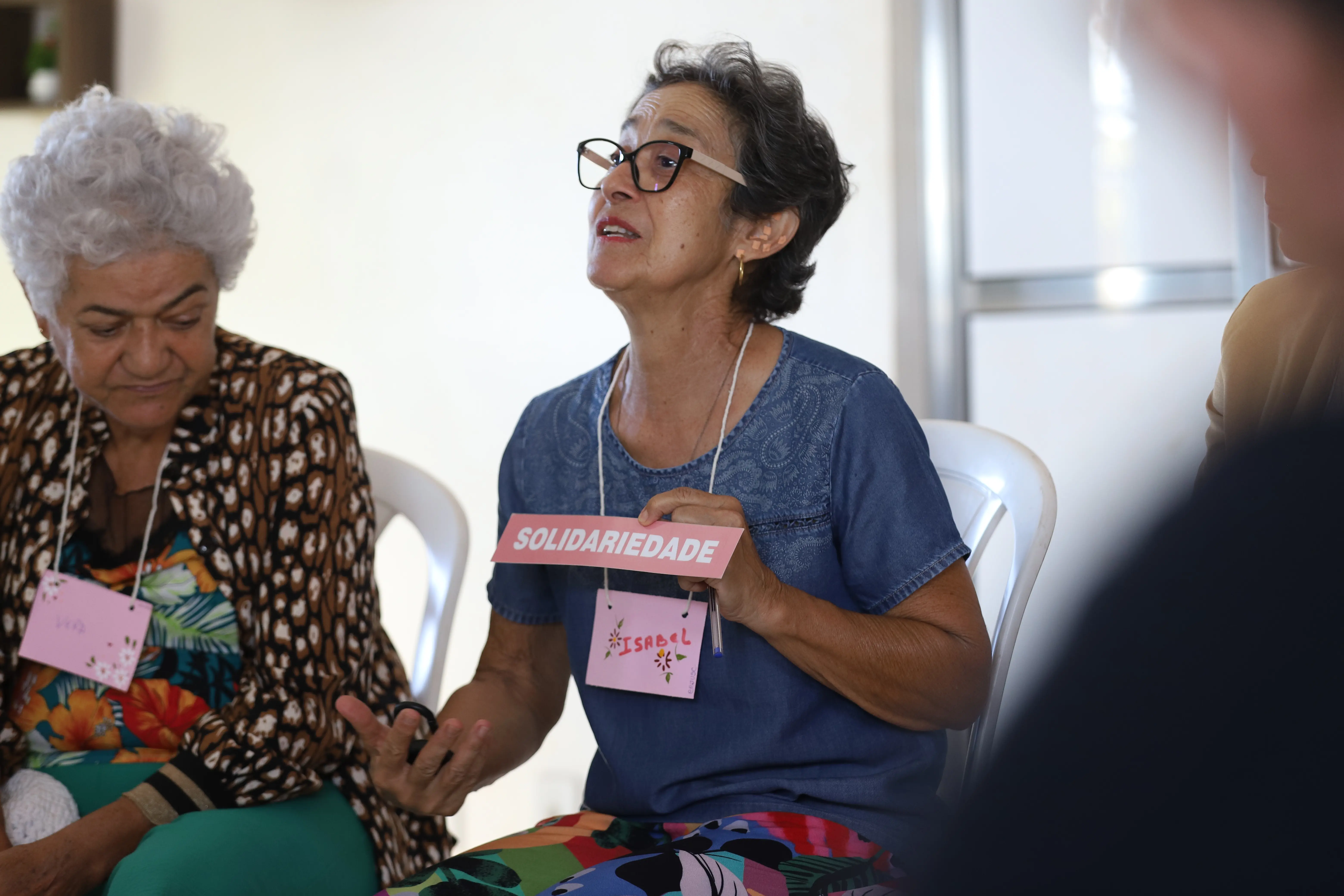 Mulher com crachá "ISABEL" compartilha a palavra “SOLIDARIEDADE” durante roda de conversa. Ela fala com emoção, destacando a importância do tema. Outra participante observa atentamente ao lado.