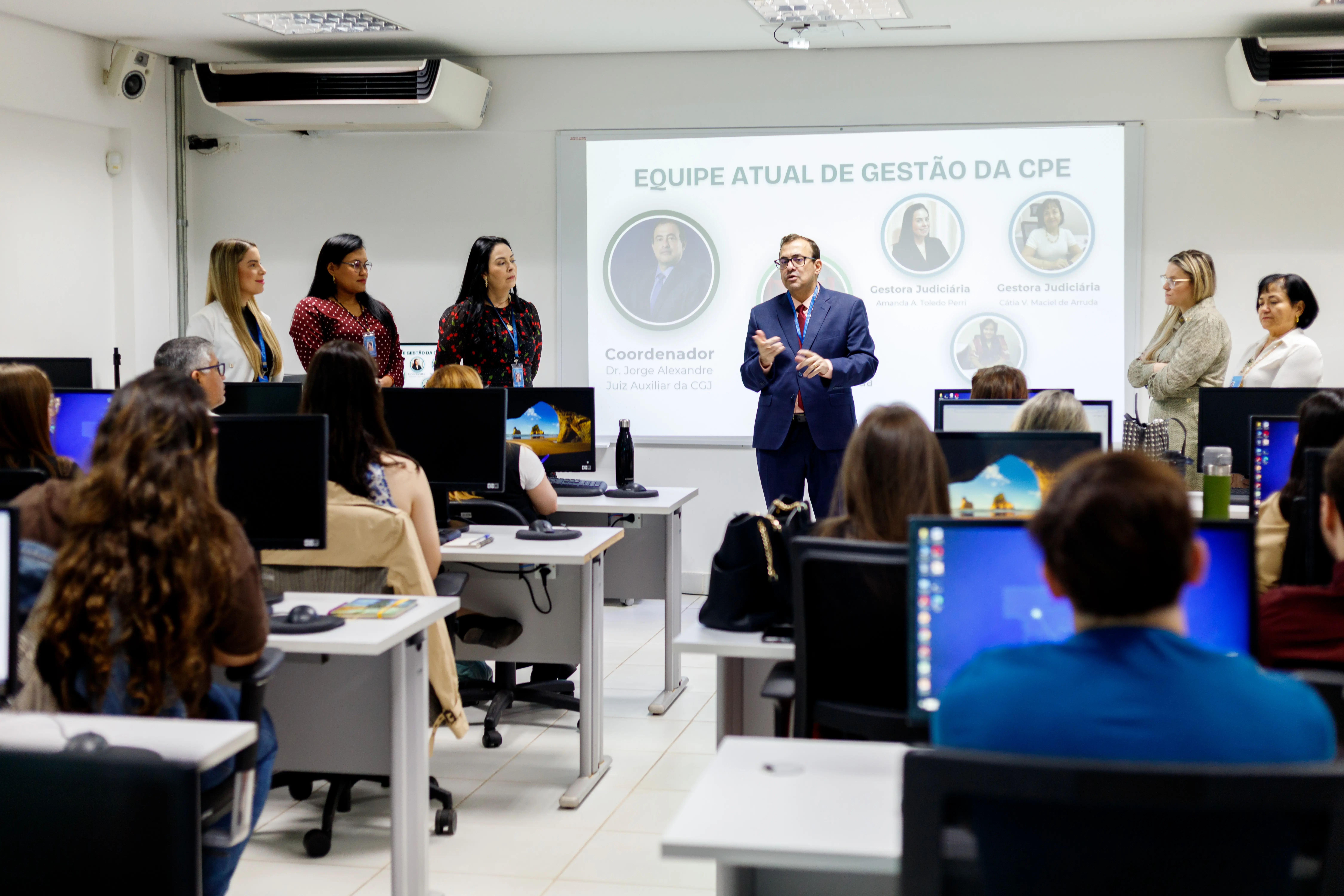 Sala de aula com computadores e telão exibe equipe de gestão da CPE; homem de terno fala ao público enquanto seis pessoas estão alinhadas à frente.