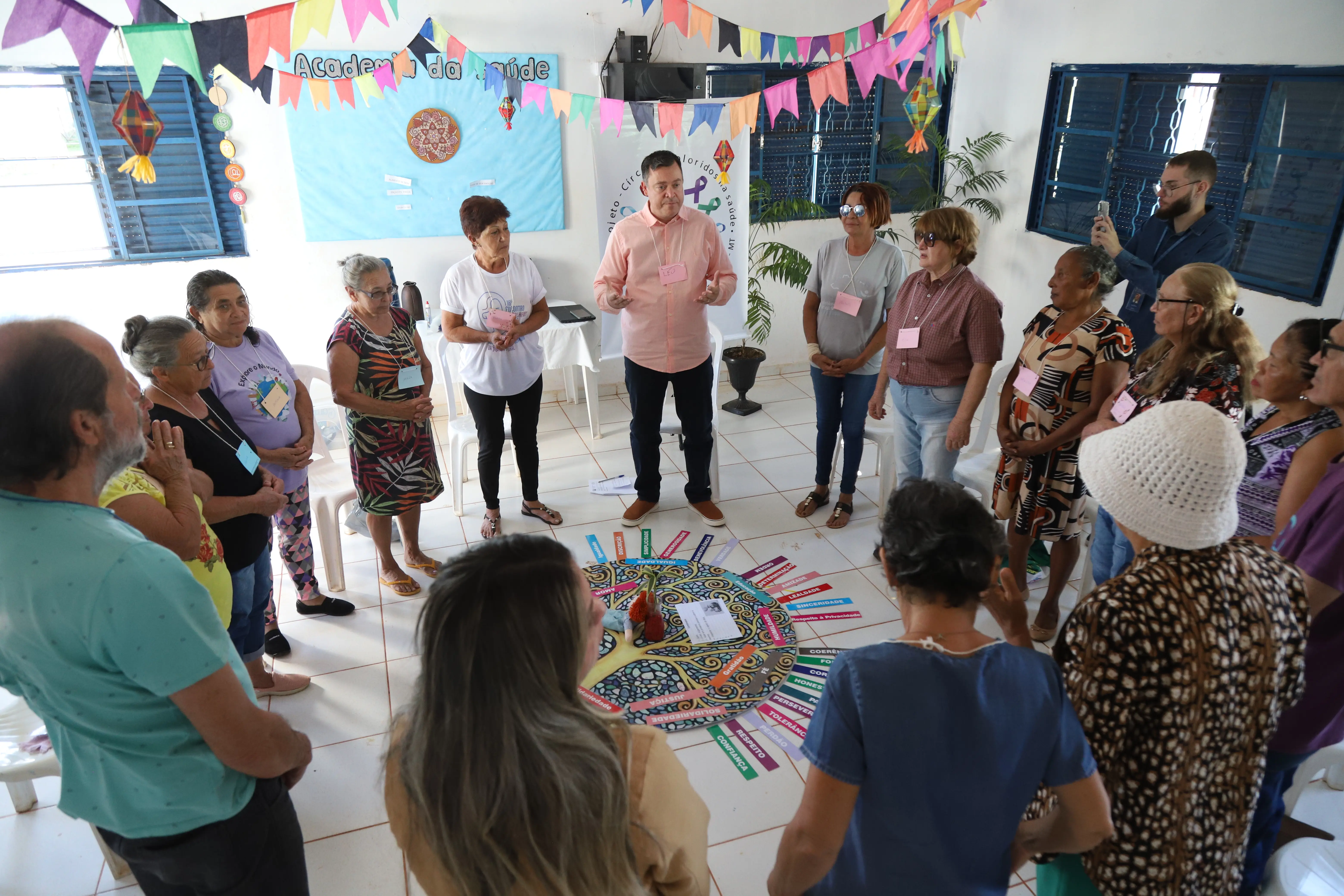 Participantes do Círculo de Construção de Paz formam uma roda em pé ao redor de mandala com palavras de valores. O facilitador conduz a atividade em ambiente decorado com bandeirolas e cartazes.