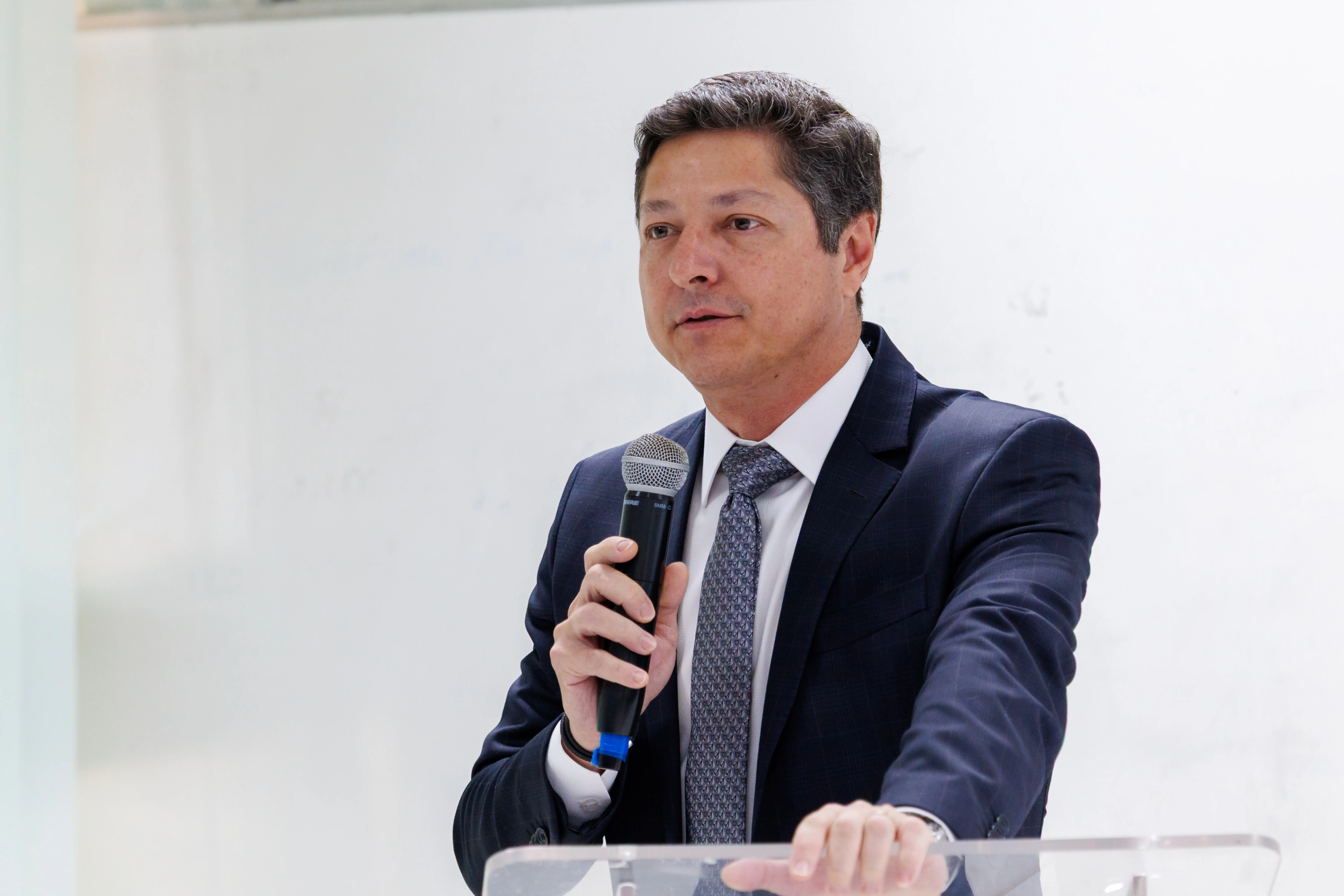 Desembargador Lídio Modesto. Homem branco, com terno preto, camisa branca, em sala com luz clara, segurando microfone durante abertura de capacitação sobre inteligência artificial.