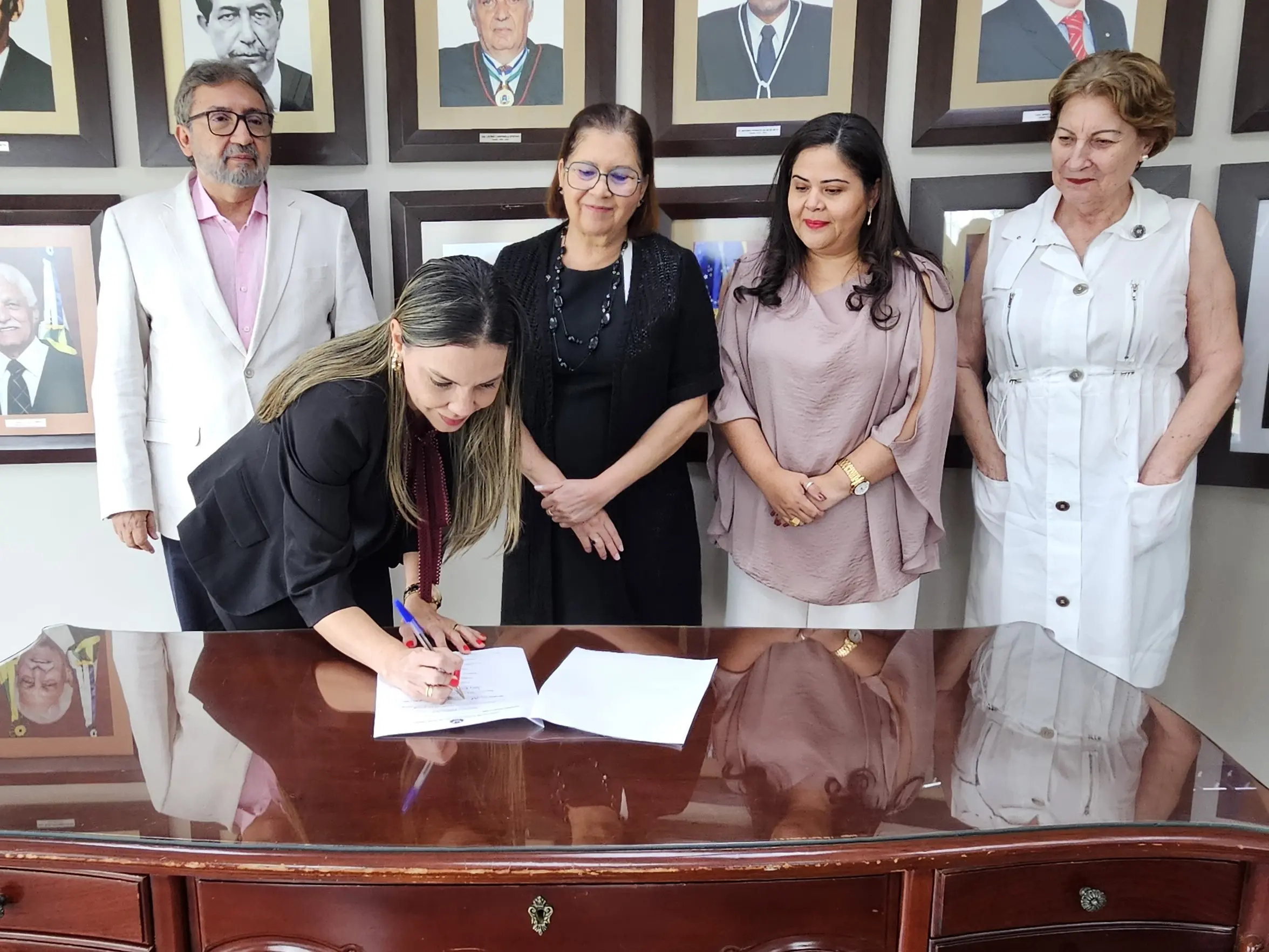 Foto colorida e horizontal. Juíza Henriqueta Lima está apoiada à mesa e assina documento de adesão. Ele veste paletó preto e calça preta. Atrás, três mulheres e um homem olham ele assinar o documento e sorriem.