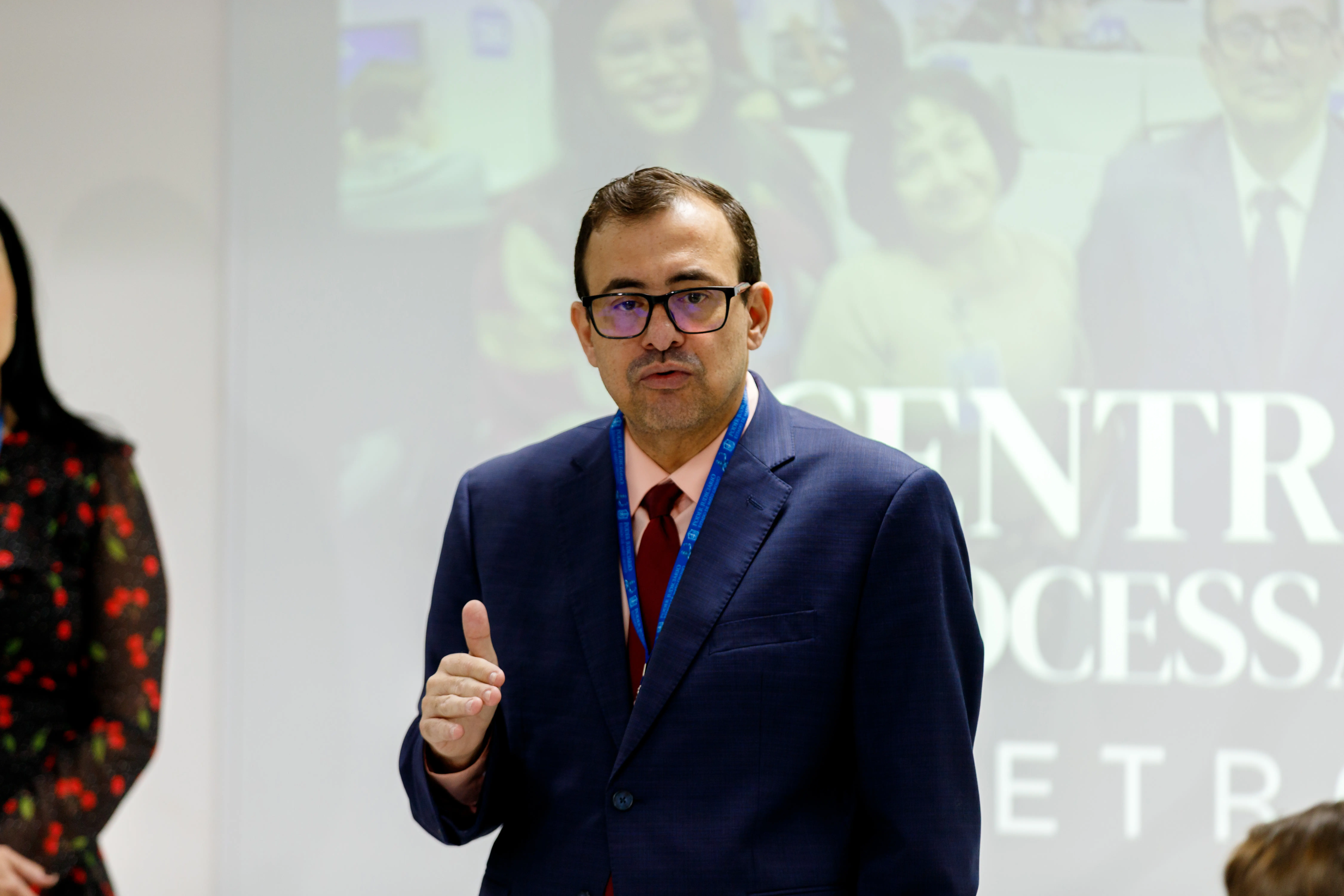 Homem de óculos, terno azul e gravata vermelha fala gesticulando em ambiente interno, com projeção ao fundo e pessoa parcialmente visível à esquerda.