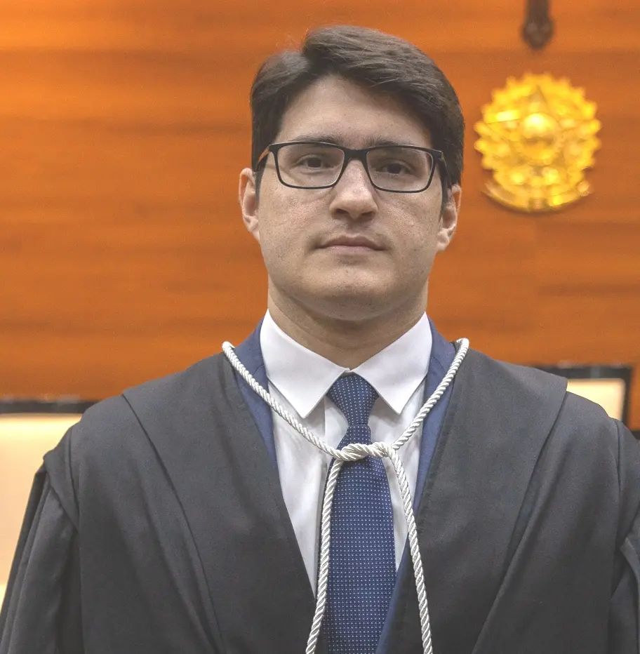 Homem de expressão séria veste toga e gravata azul com fundo pontilhado. Ao fundo, o brasão da República indica ambiente solene, possivelmente uma cerimônia no Poder Judiciário. 