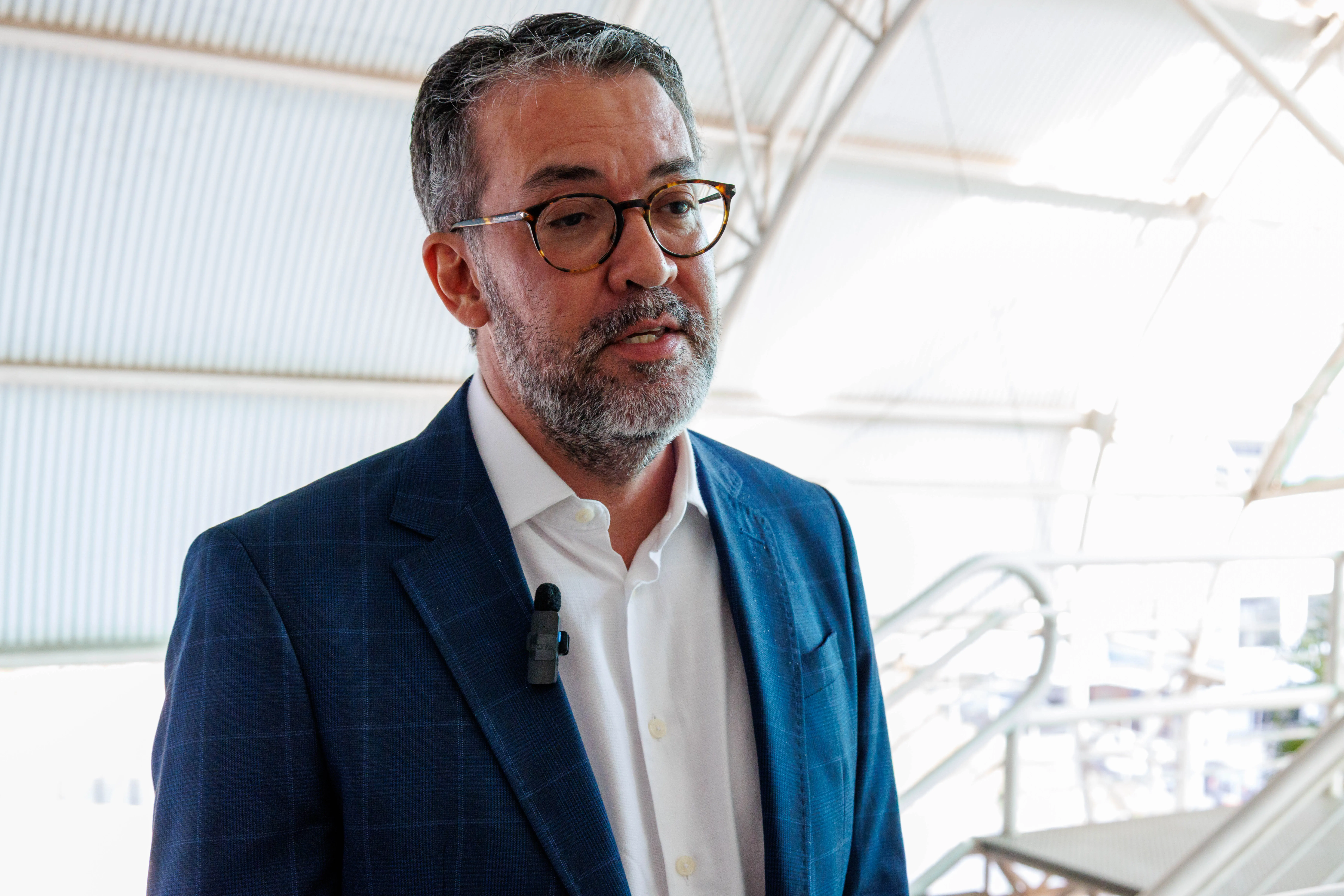 defensor público Alison Costa Ourives é um homem de terno azul fala com microfone preso à lapela, em ambiente iluminado.