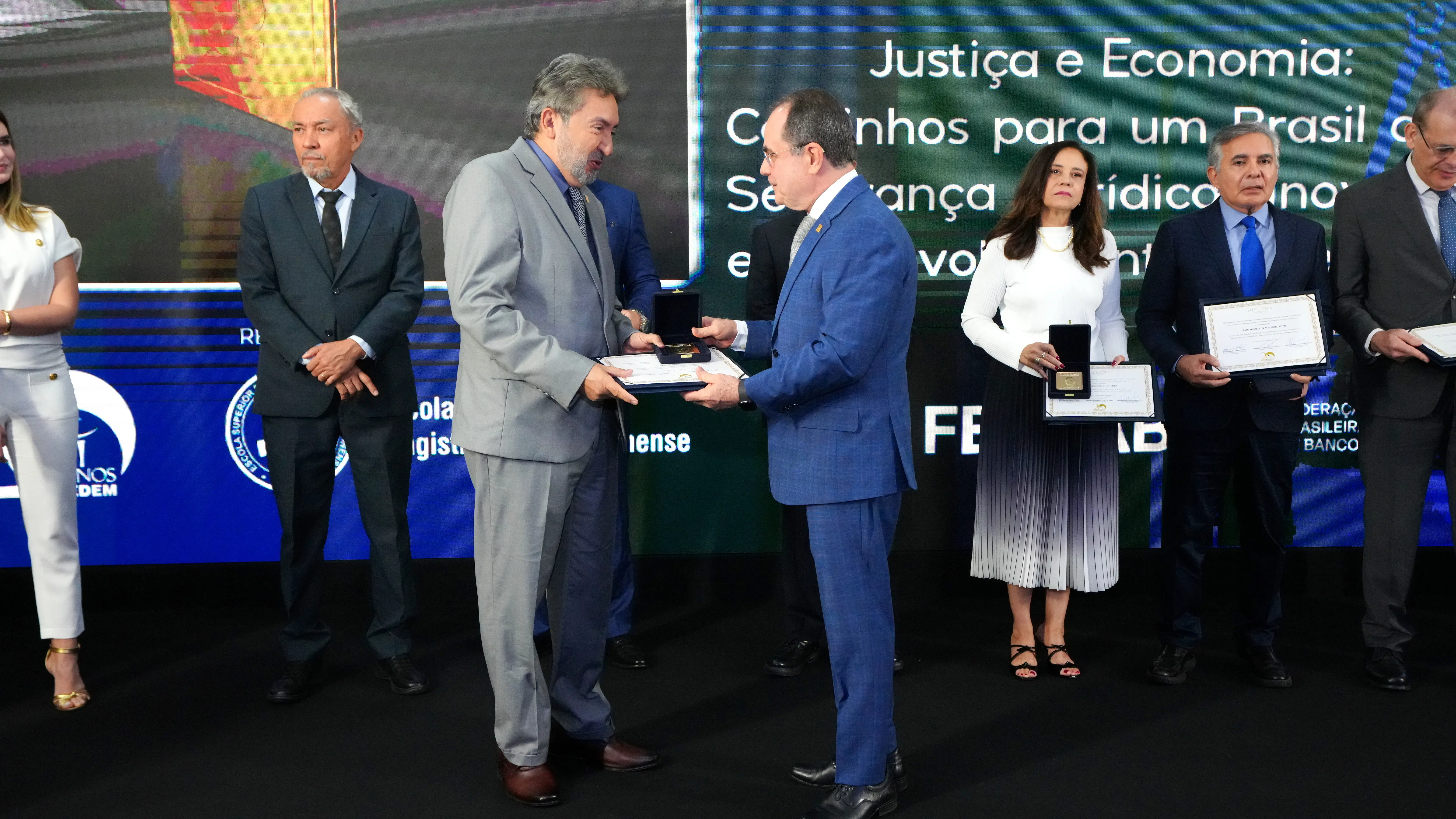 fotografia colorida do des. Márcio Vidal (homem branco, de cabelos e barba grisalhos, e roupa social cinza) recebendo a homenagem as mãos do des. Marco Villas Boas, que é um homem branco, de óculos de grau, que usa roupa social azul. 