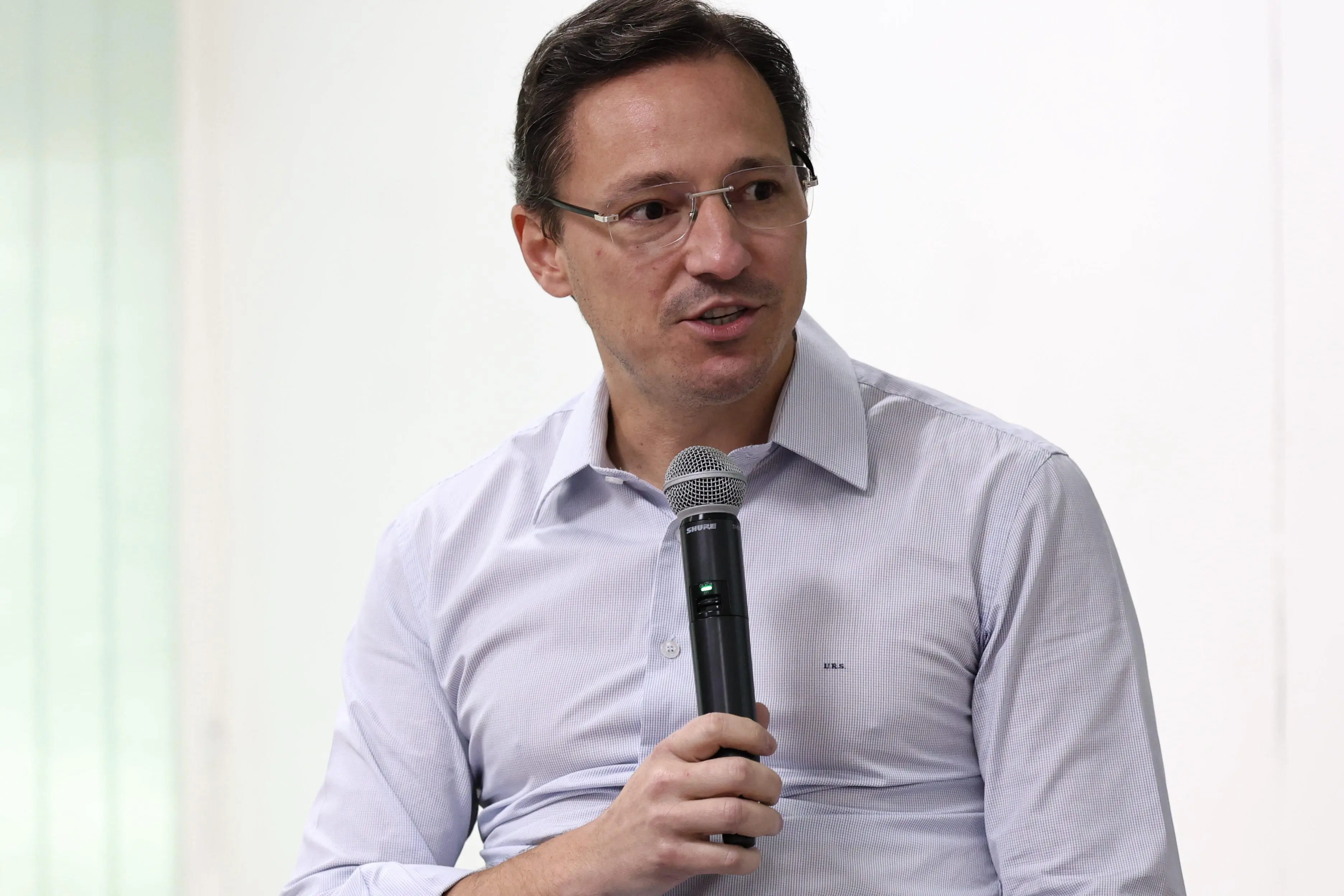 Ulisses Rabaneda é um homem branco, usa óculos de grau, camisa clara e fala ao microfone