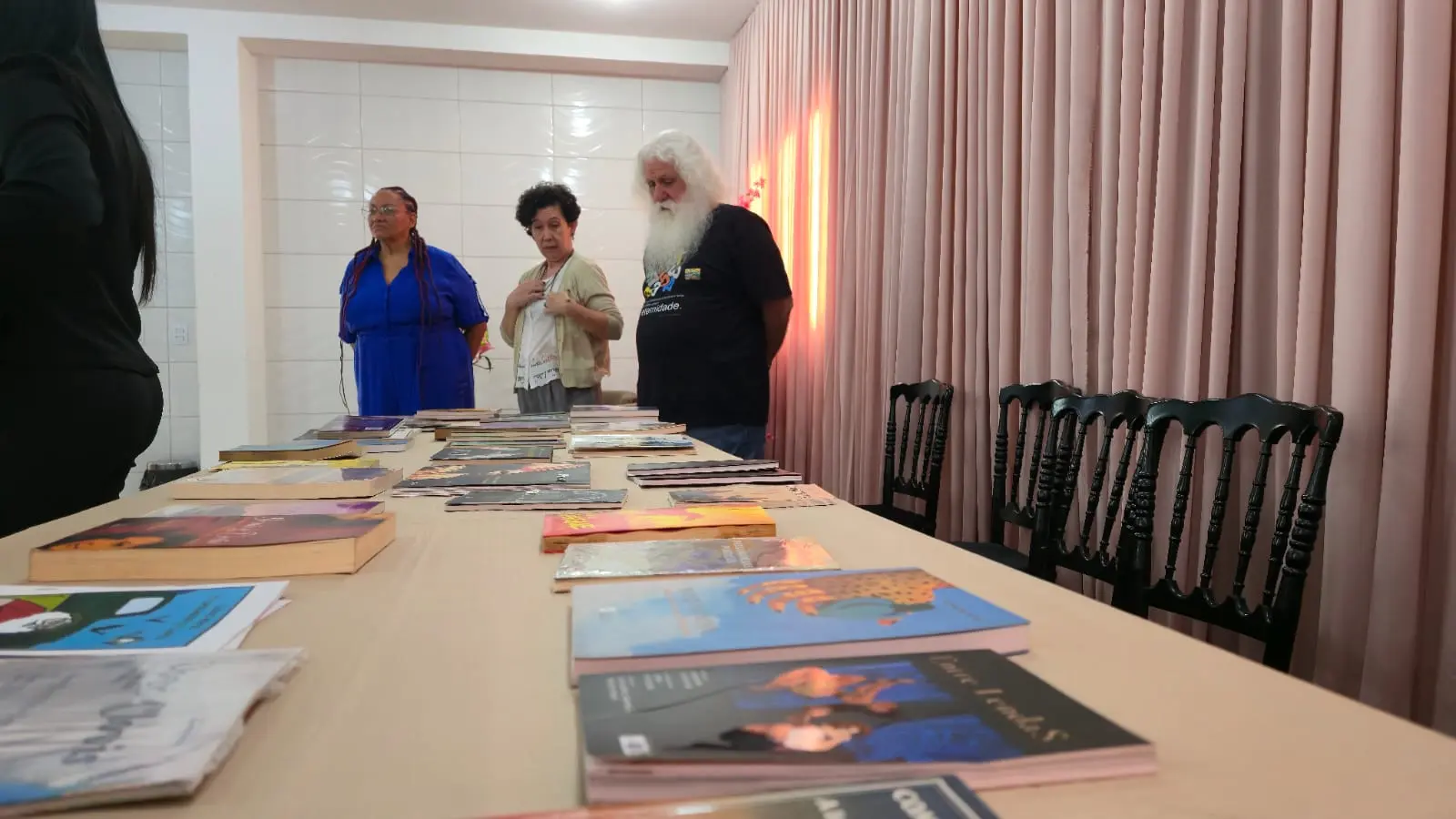 Três pessoas estão atrás de uma mesa longa com vários livros dispostos. Da esquerda para a direita, uma mulher negra com roupa azul, uma mulher de cabelo cacheado curto e um homem com cabelos e barba longos e brancos. No fundo, uma cortina de cor de rosa.