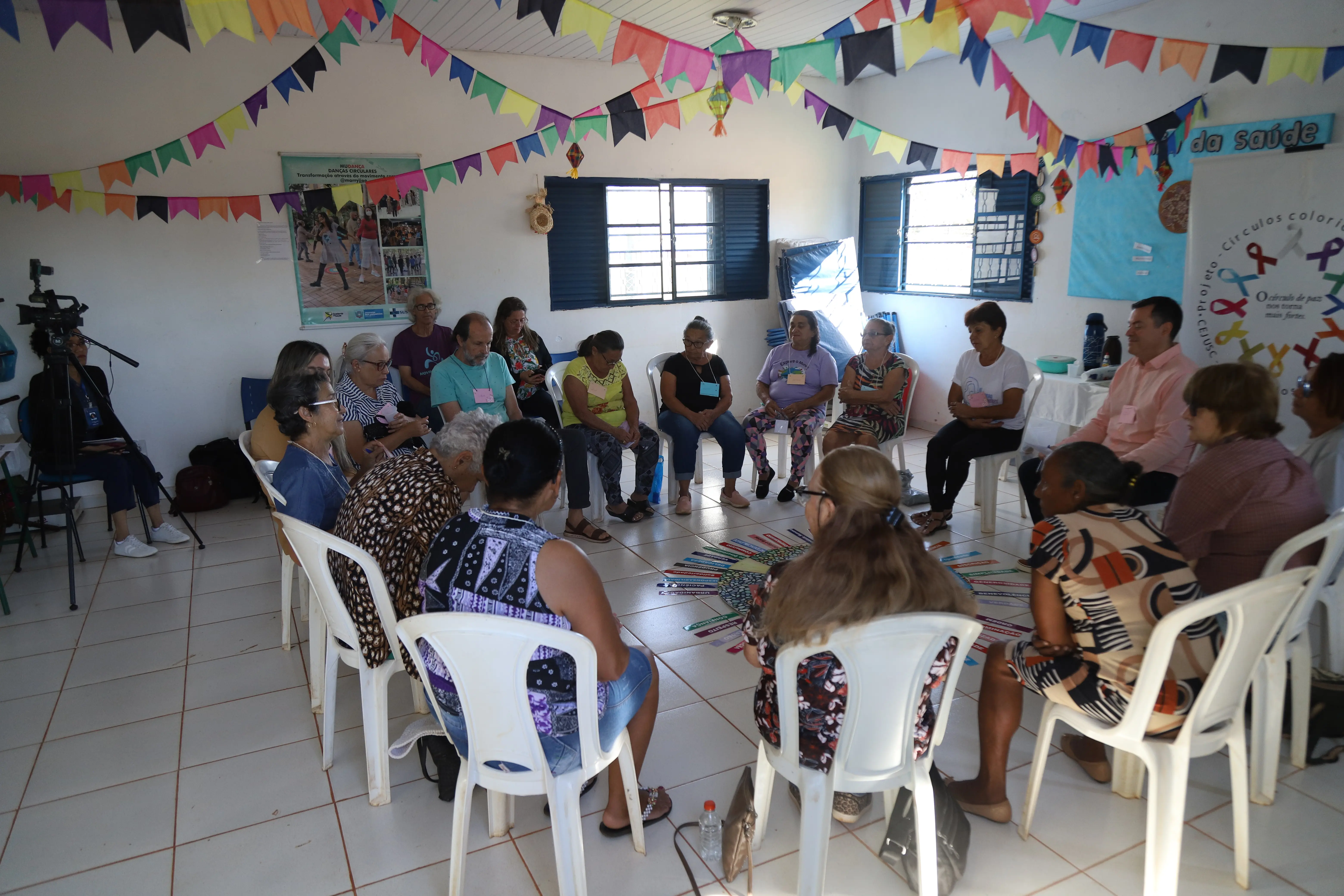 Grupo de idosos e facilitadores senta-se em roda ao redor de mandala com palavras como “amizade” e “respeito”. O ambiente decorado com bandeirinhas e câmeras sugere atividade acompanhada por mídia ou registro institucional.