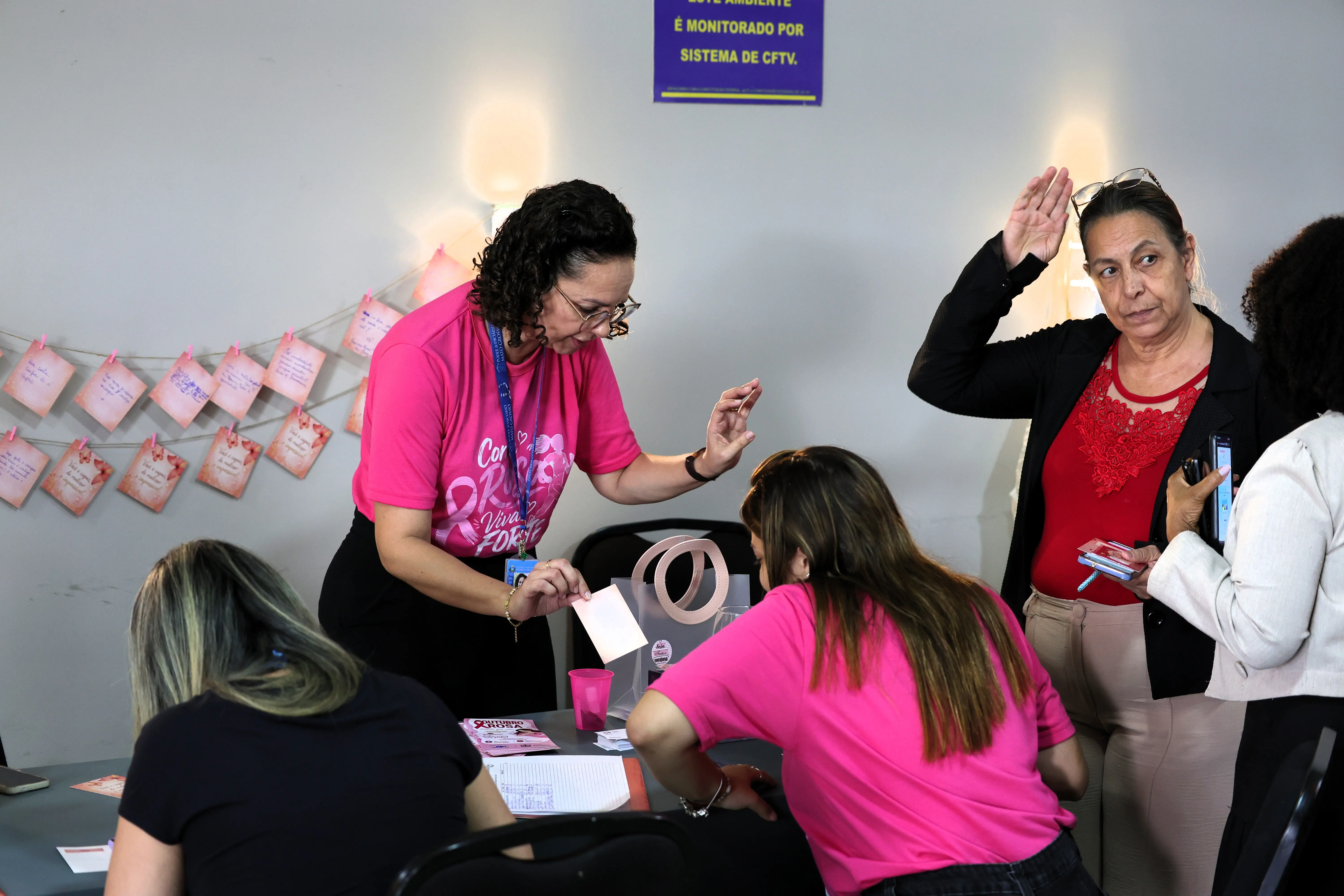 Mulher de cabelos cacheados, óculos e camiseta rosa entrega um cartão a participante sentada à mesa; ao fundo, mensagens em papéis rosa presos a um varal.