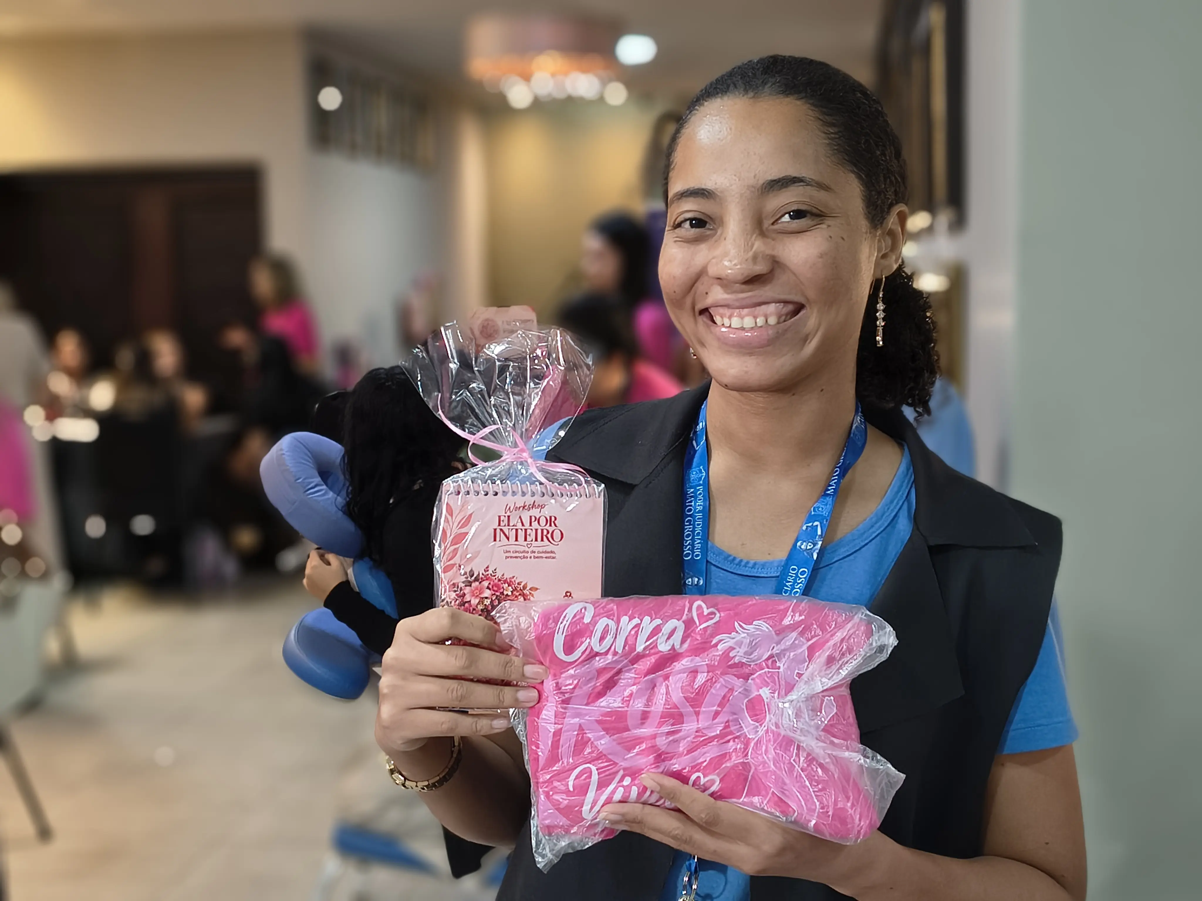 Mulher de cabelo preso e colete preto sorri e segura o caderno do workshop e uma camiseta rosa, recebidos no evento; ao fundo, participantes.