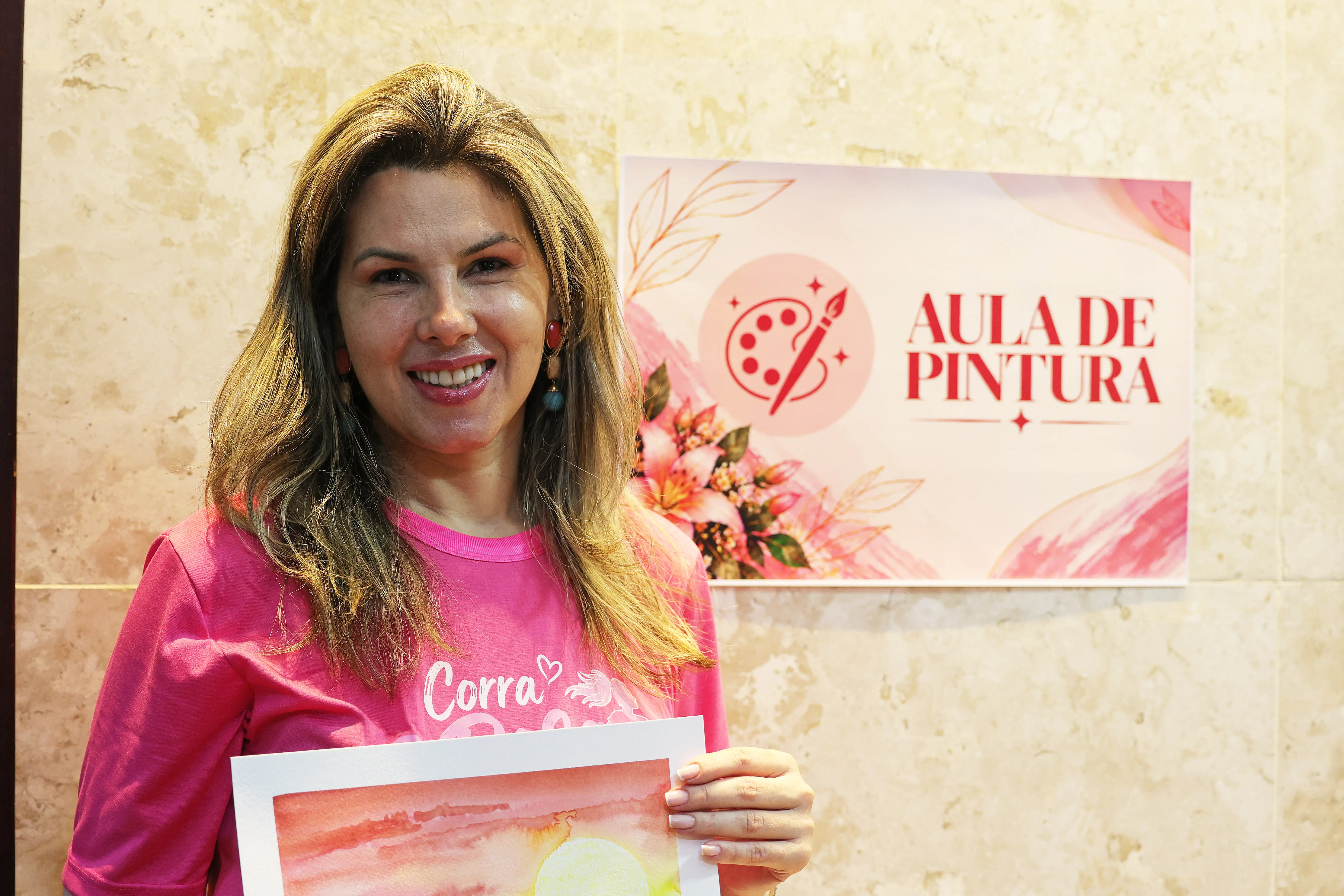 Mulher de cabelos castanhos e camiseta rosa sorri e segura uma pintura em aquarela de pôr do sol, ao lado do cartaz da aula de pintura.