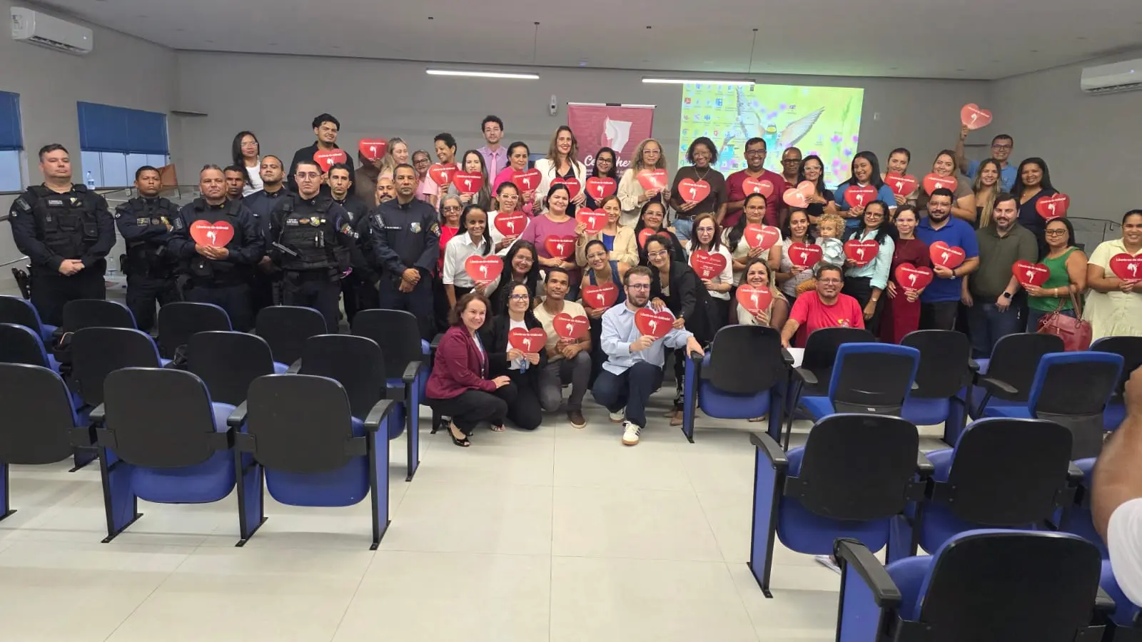 Foto horizontal que mostra um auditório e dezenas de pessoas em pé, posando para a foto, segurando panfletos vermelhos em formato de coração.